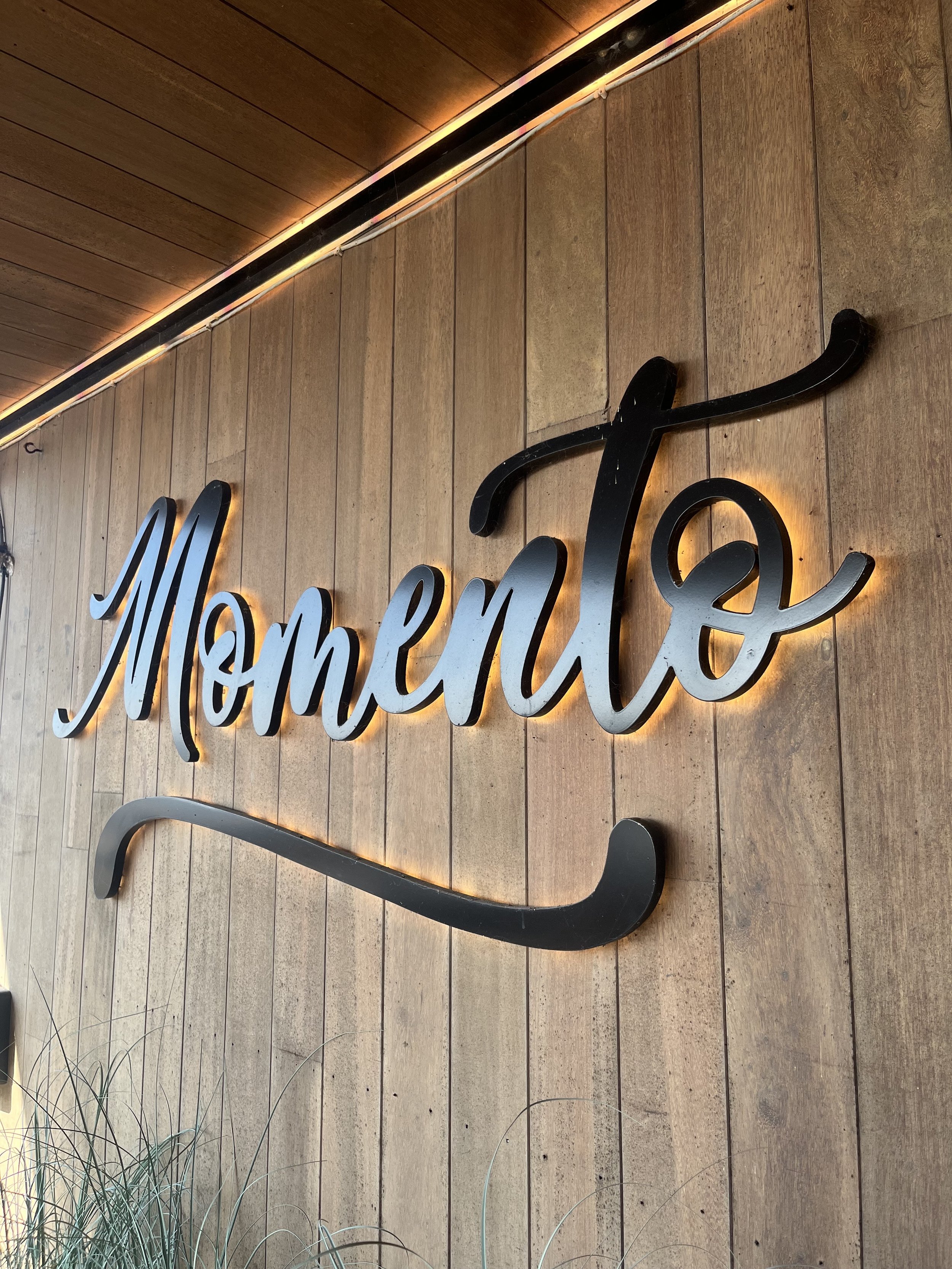 Houten muur met het woord 'Momento' in sierlijke letters bevestigd, verlicht door zachte verlichting.