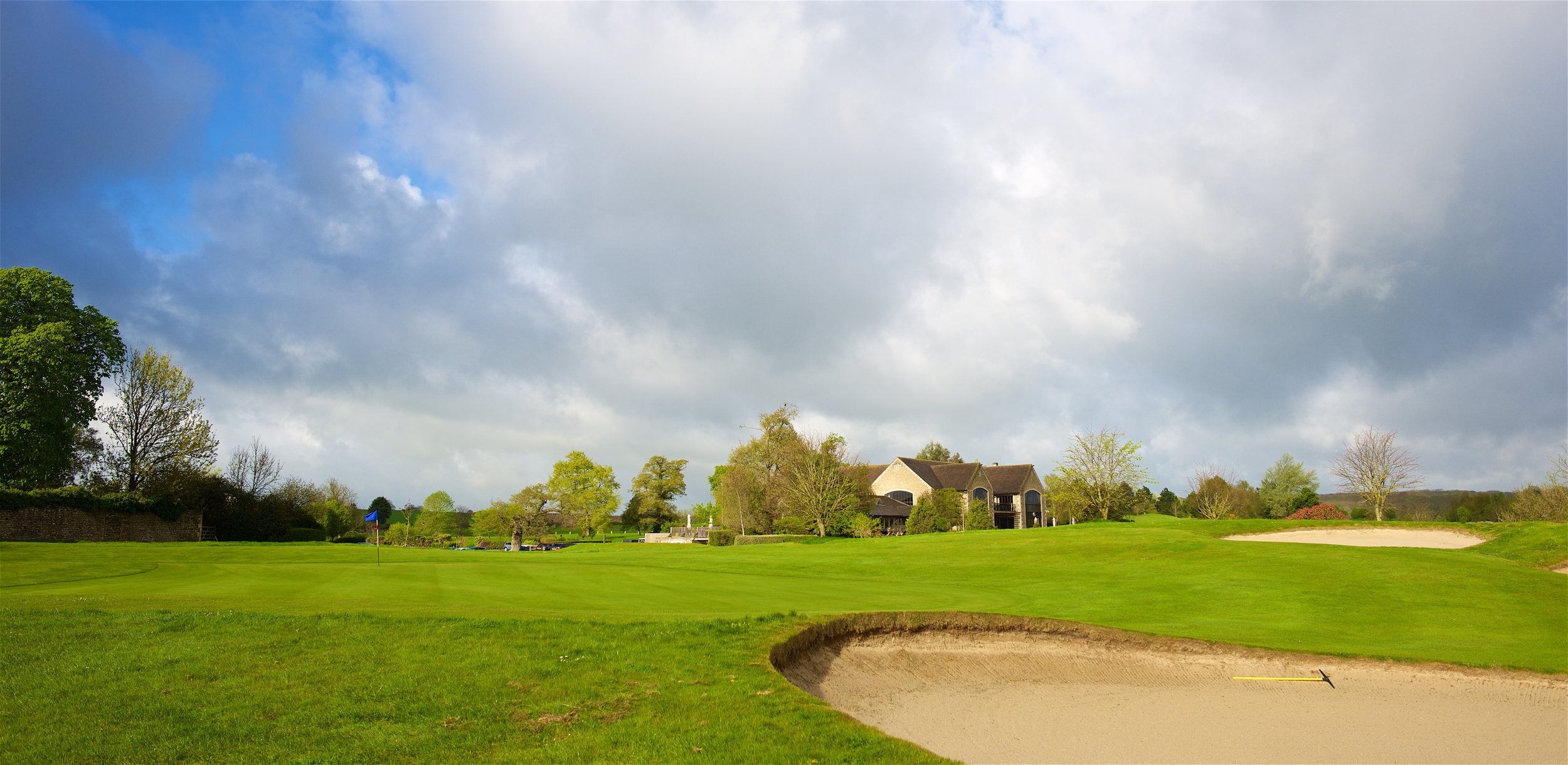 A golf course with a sand bunker in the foreground, lush green grass, trees, a building in the distance, and a partly cloudy sky.