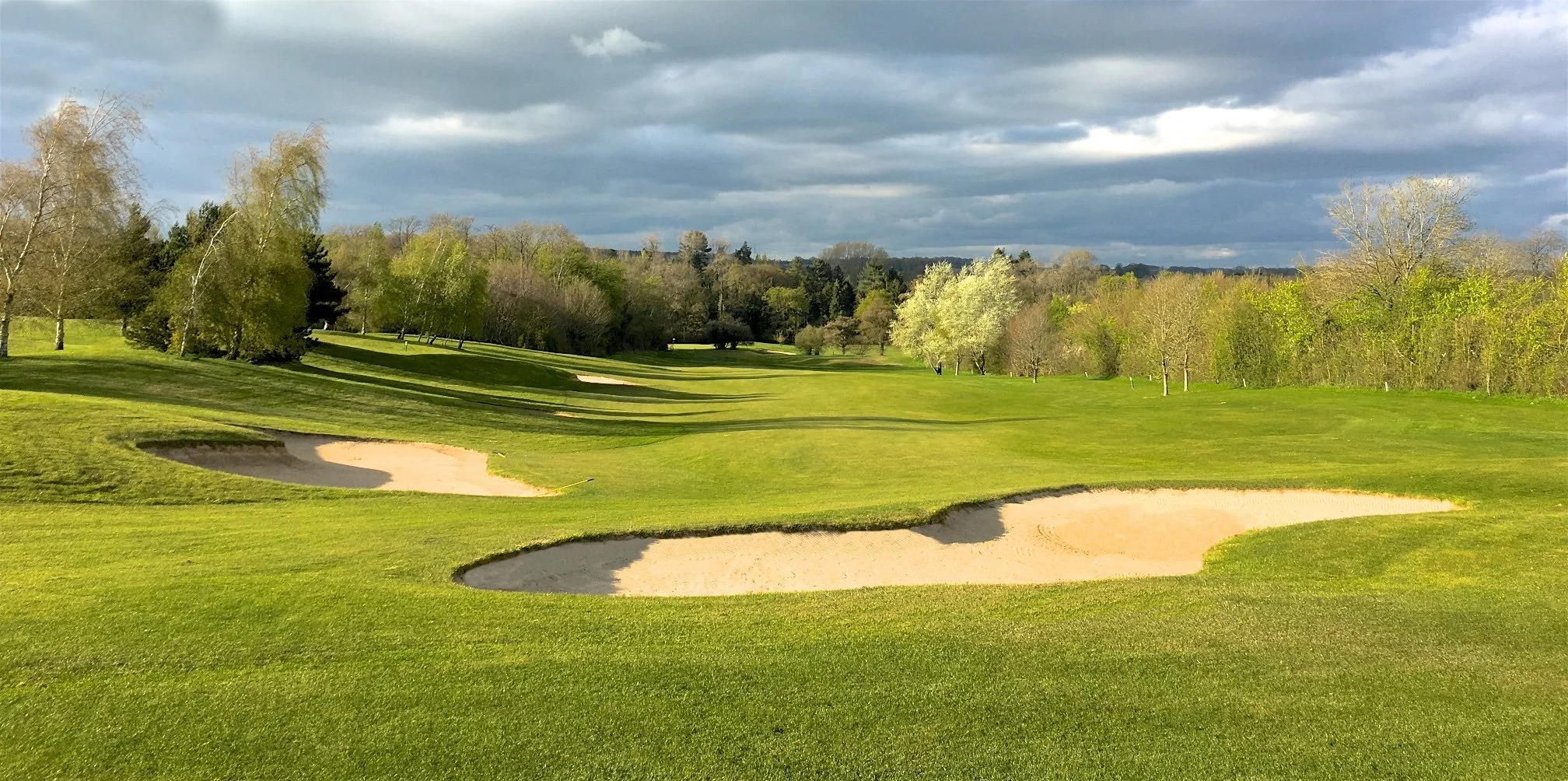 A scenic golf course view with lush green fairways, sand bunkers, trees with budding leaves, and a cloudy sky overhead.