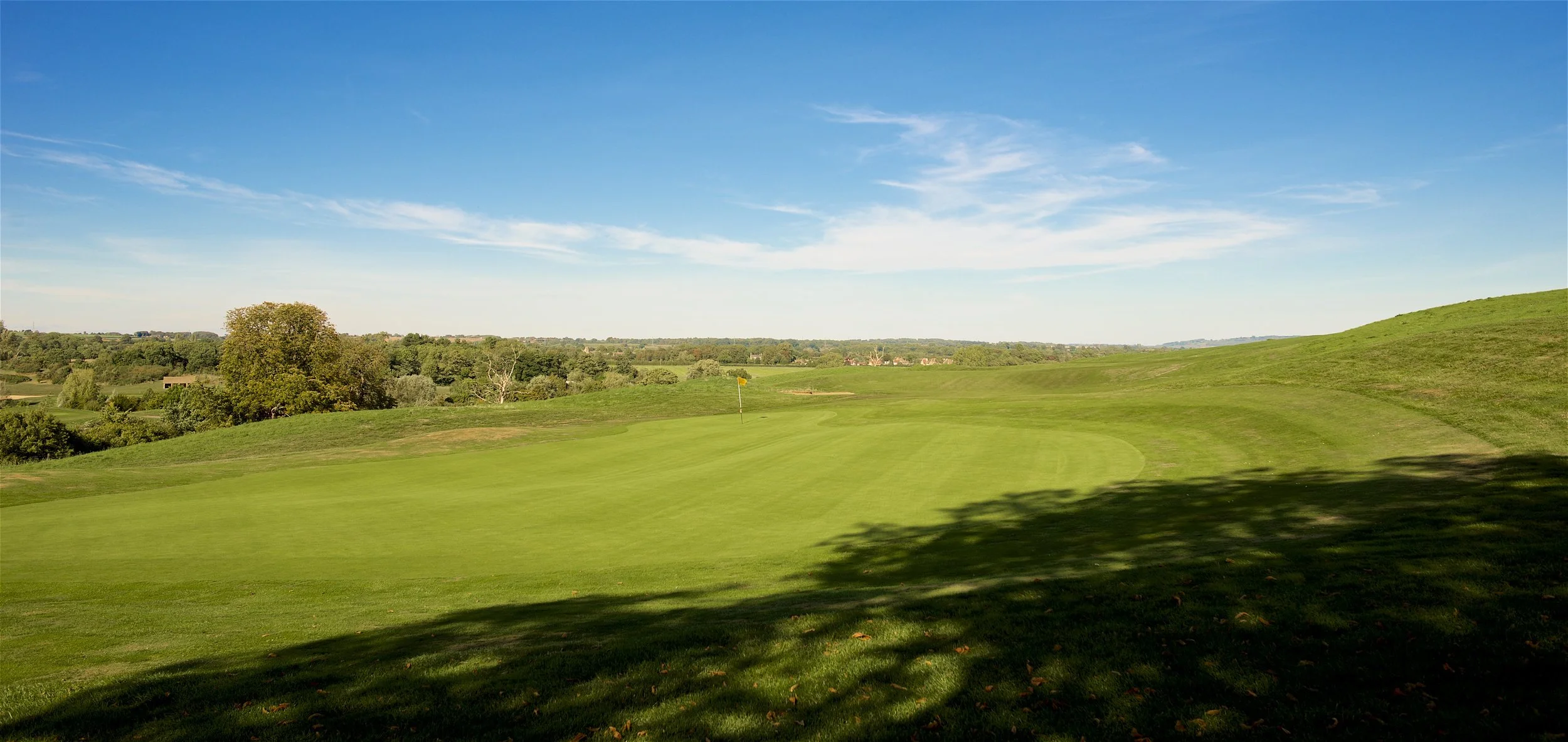 A golf course with a green putting area, a yellow flag marking the hole, surrounded by rolling hills, trees, and a clear blue sky.