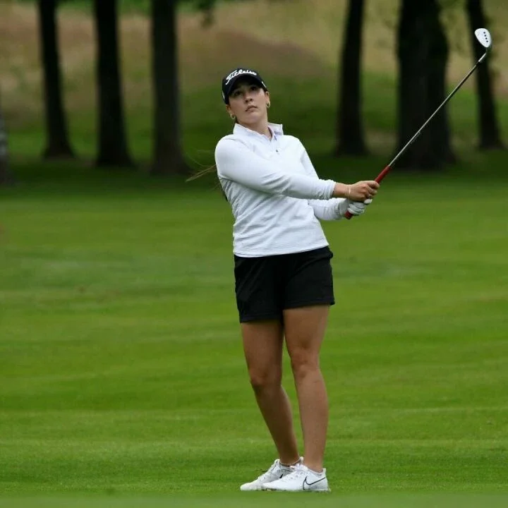 A woman playing golf on a green golf course, holding a golf club, wearing a white long-sleeve shirt, black shorts, white sneakers, and a black cap.