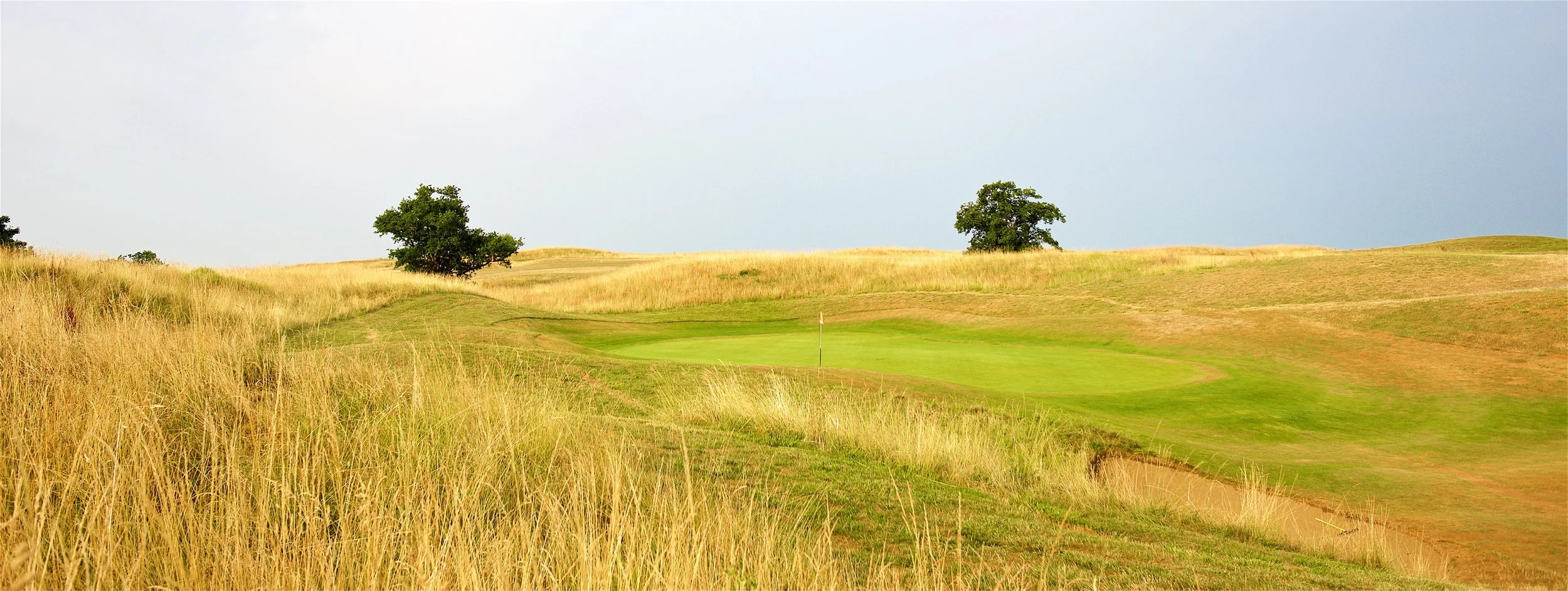 A golf course with a putting green, surrounded by grassy hills and a few trees, under a partly cloudy sky.