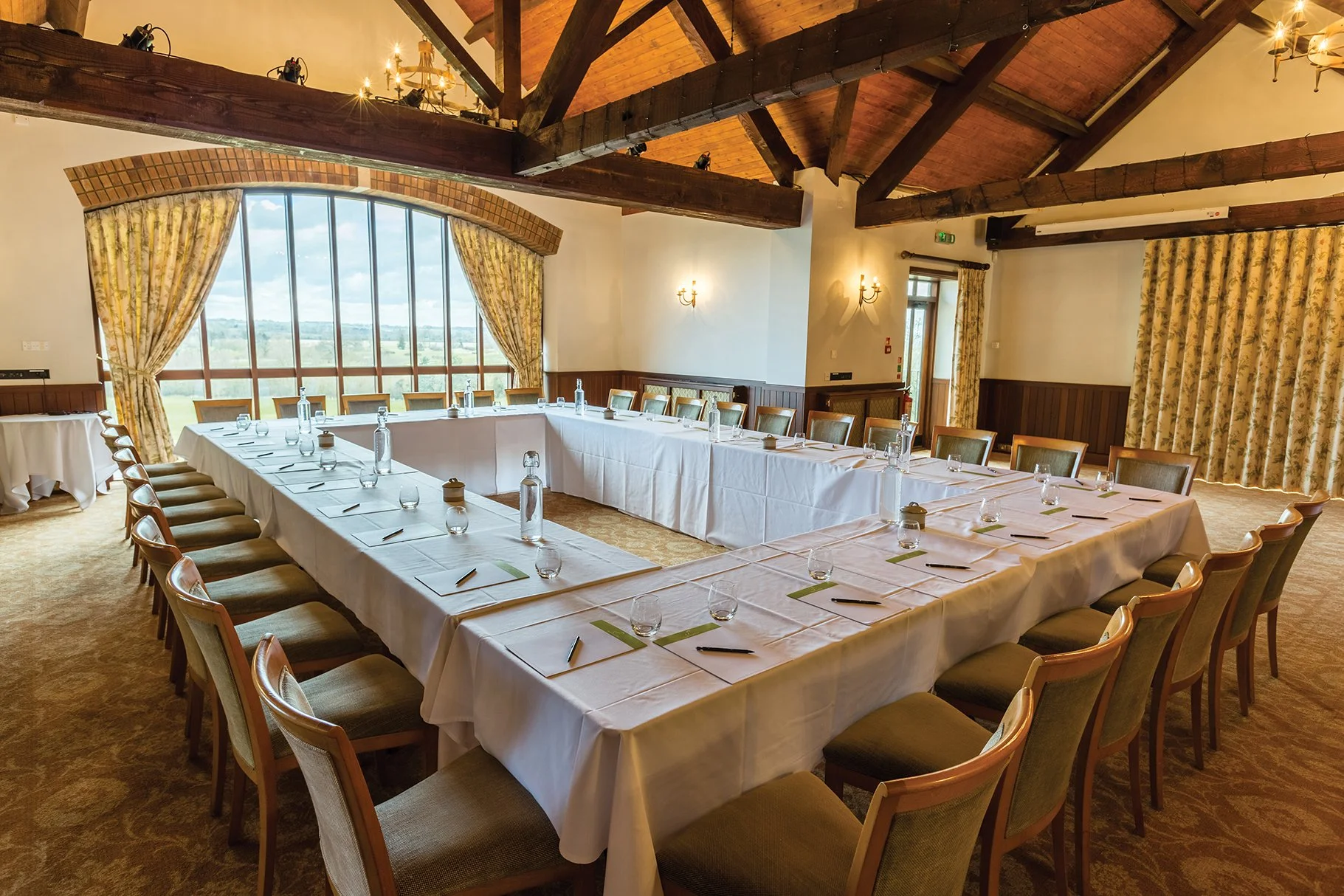 A large indoor conference room with a U-shaped arrangement of long tables covered with white tablecloths and arranged with notepads, pens, water bottles, and glasses. The room has a high vaulted wooden ceiling with exposed beams, large windows with yellow floral curtains, and a view of the outdoors. The walls have wall-mounted lamps, and there are chandeliers hanging from the ceiling.