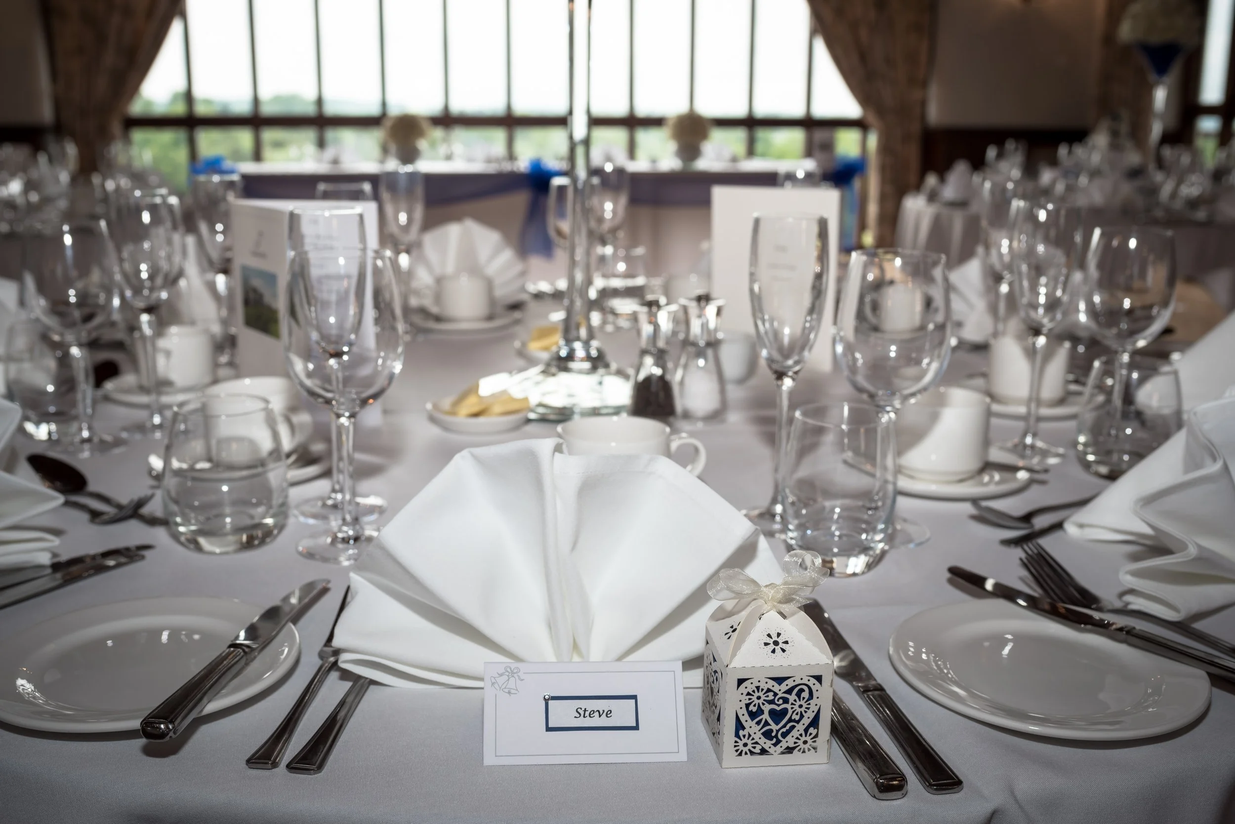 A banquet table set for a formal event with white tablecloths, multiple wine glasses, silverware, white napkins, and a decorative lantern, with a window showing greenery outside in the background.