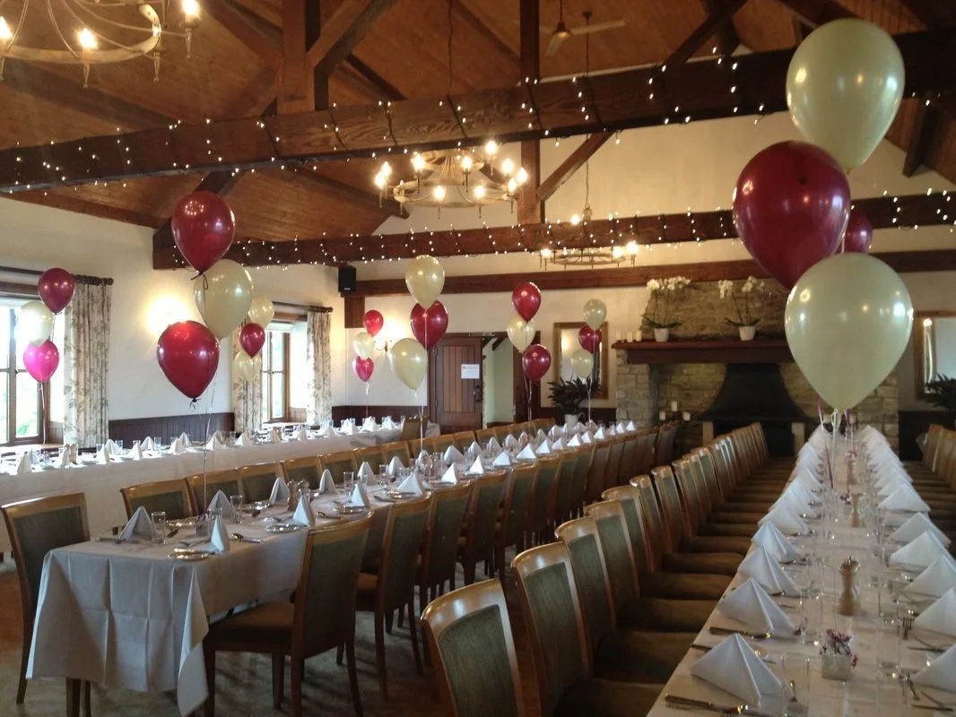 Decorated banquet hall with long tables, white tablecloths, white napkins, and balloons in maroon, pink, and cream colors. The hall features wooden beams, chandeliers, string lights, windows with floral curtains, and a brick fireplace.