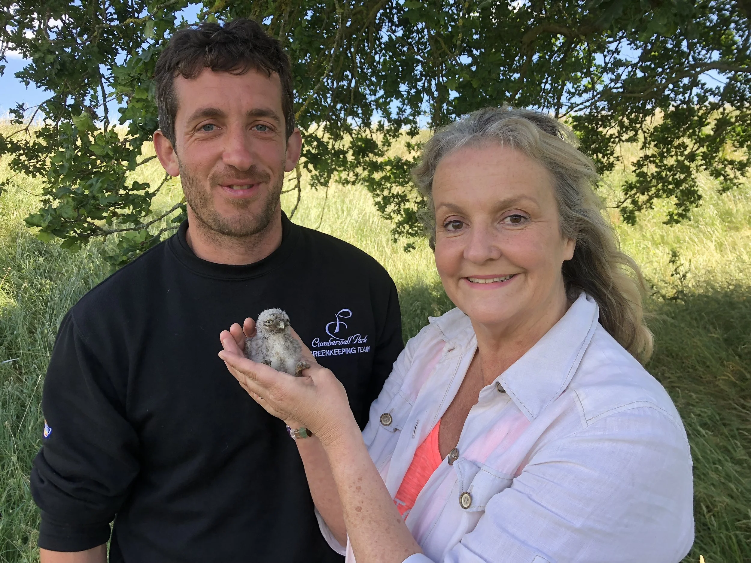 A man and a woman outdoors under a tree, holding a small bird. The man is wearing a black shirt with a logo, and the woman is wearing a light-colored shirt. They are smiling at the camera.