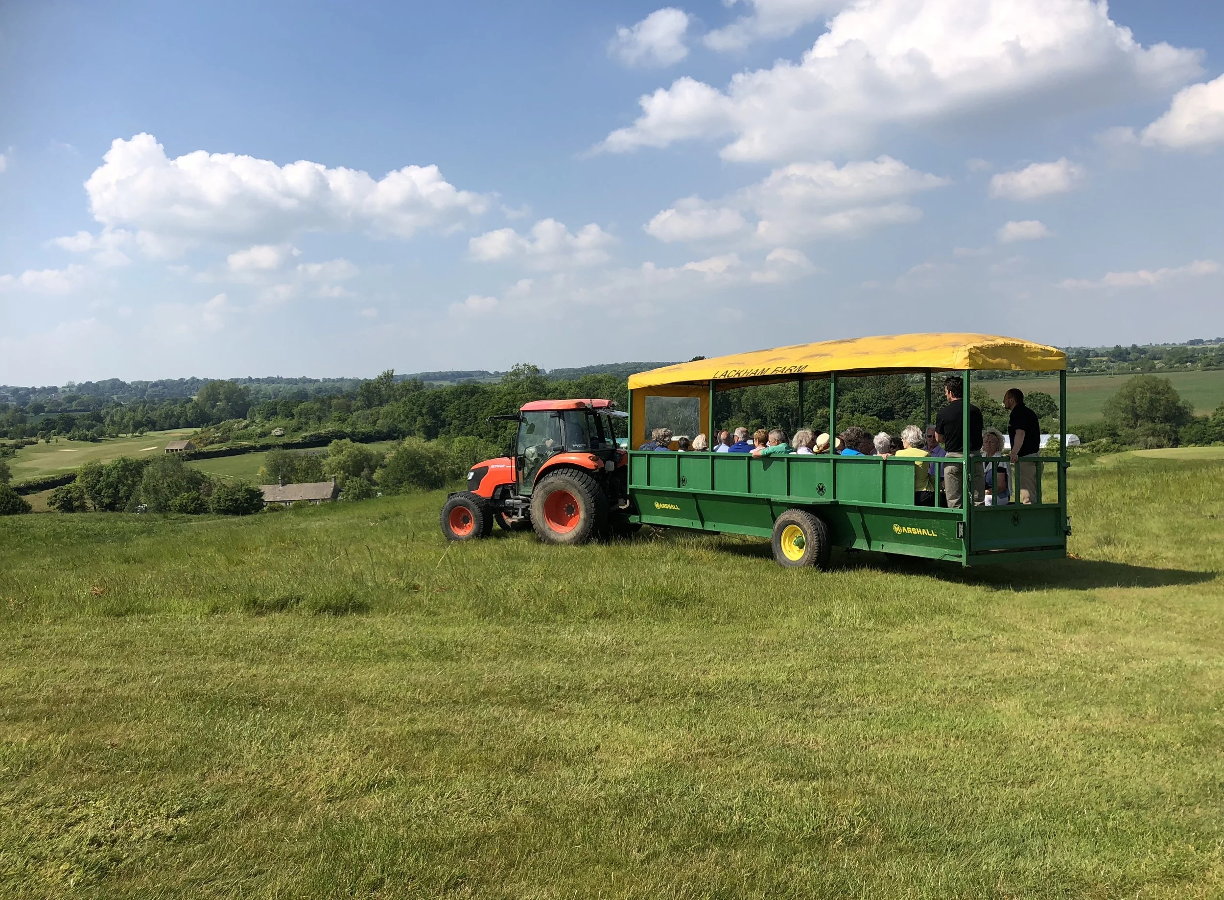 A group of people on a tractor-pulled wagon with a yellow canopy labeled 'LACKHAM FARM' in a rural landscape with green fields and trees under a blue sky with scattered clouds.
