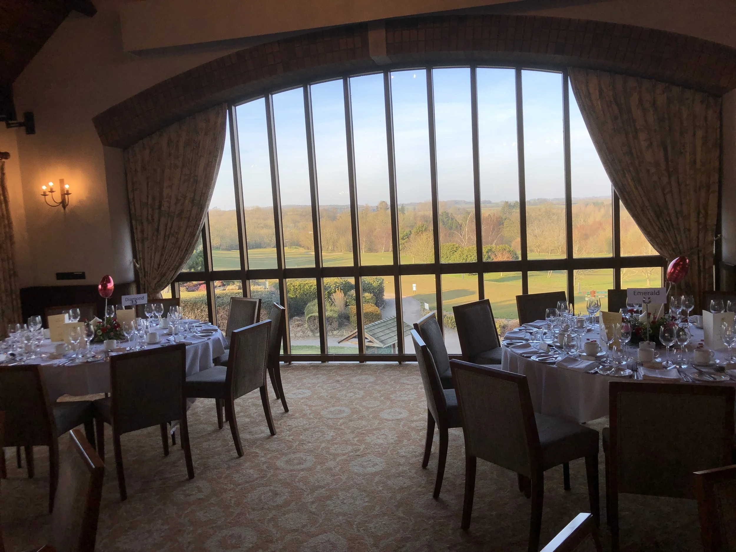 Indoor banquet hall with large glass window showing a view of a field and trees outside. The room is decorated for a formal event with round tables, white tablecloths, and many wine glasses. There are name cards on the tables, and a floral centerpiec
