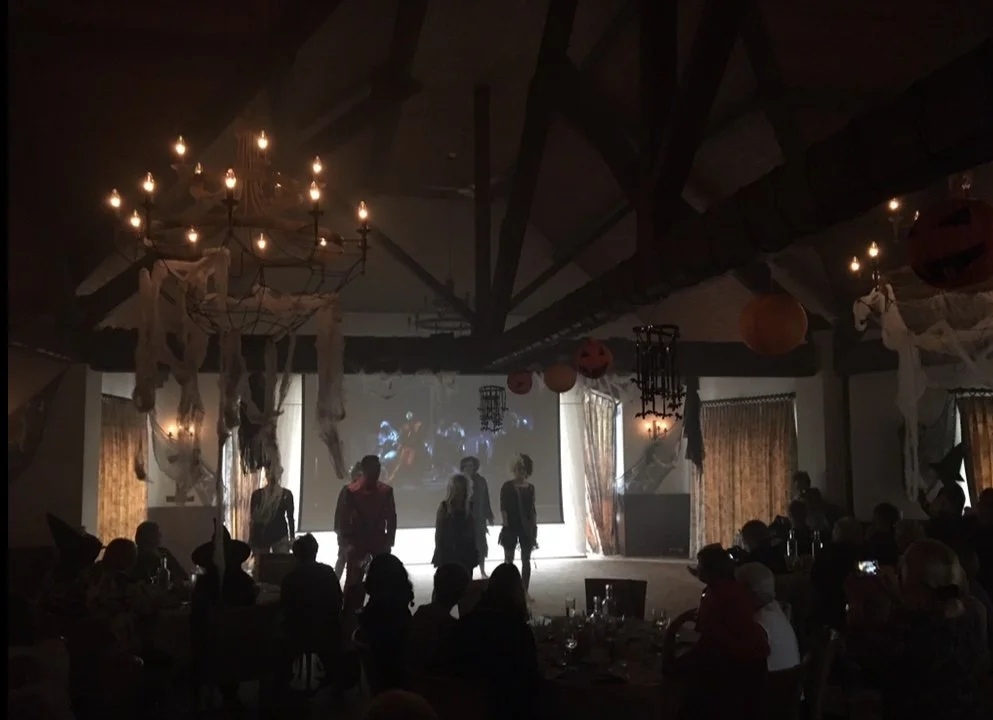 A Halloween-themed party scene with dim lighting, decorated with hanging fake spider webs, pumpkins, and orange lanterns, featuring a group of people on a stage and an audience seated at tables.