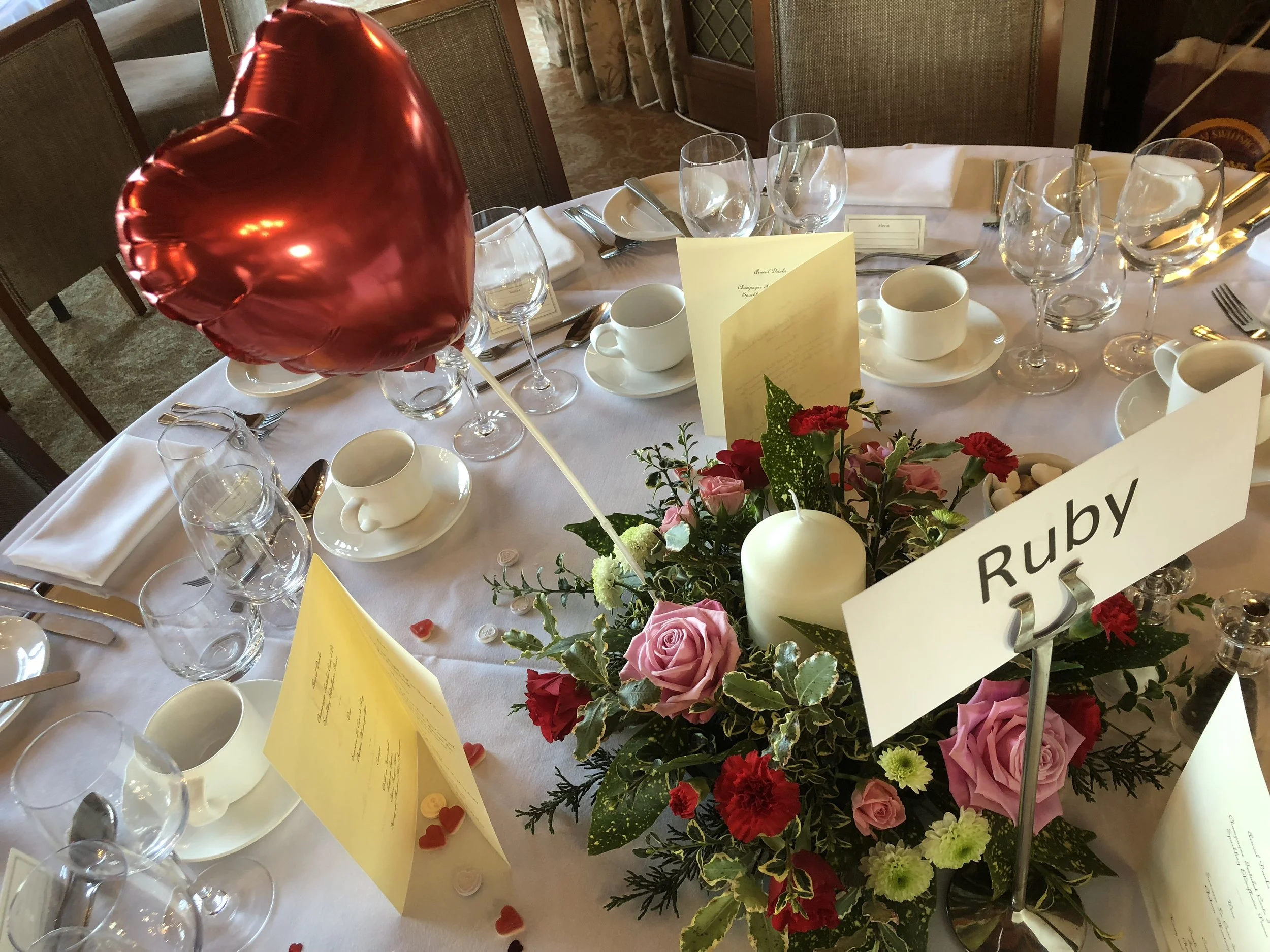 A table set for a celebration with glasses, cups, and silverware, decorated with a bouquet of pink, red, and green flowers, with a bright red heart-shaped balloon anchored to it, and a yellow paper menu or place card on the table.