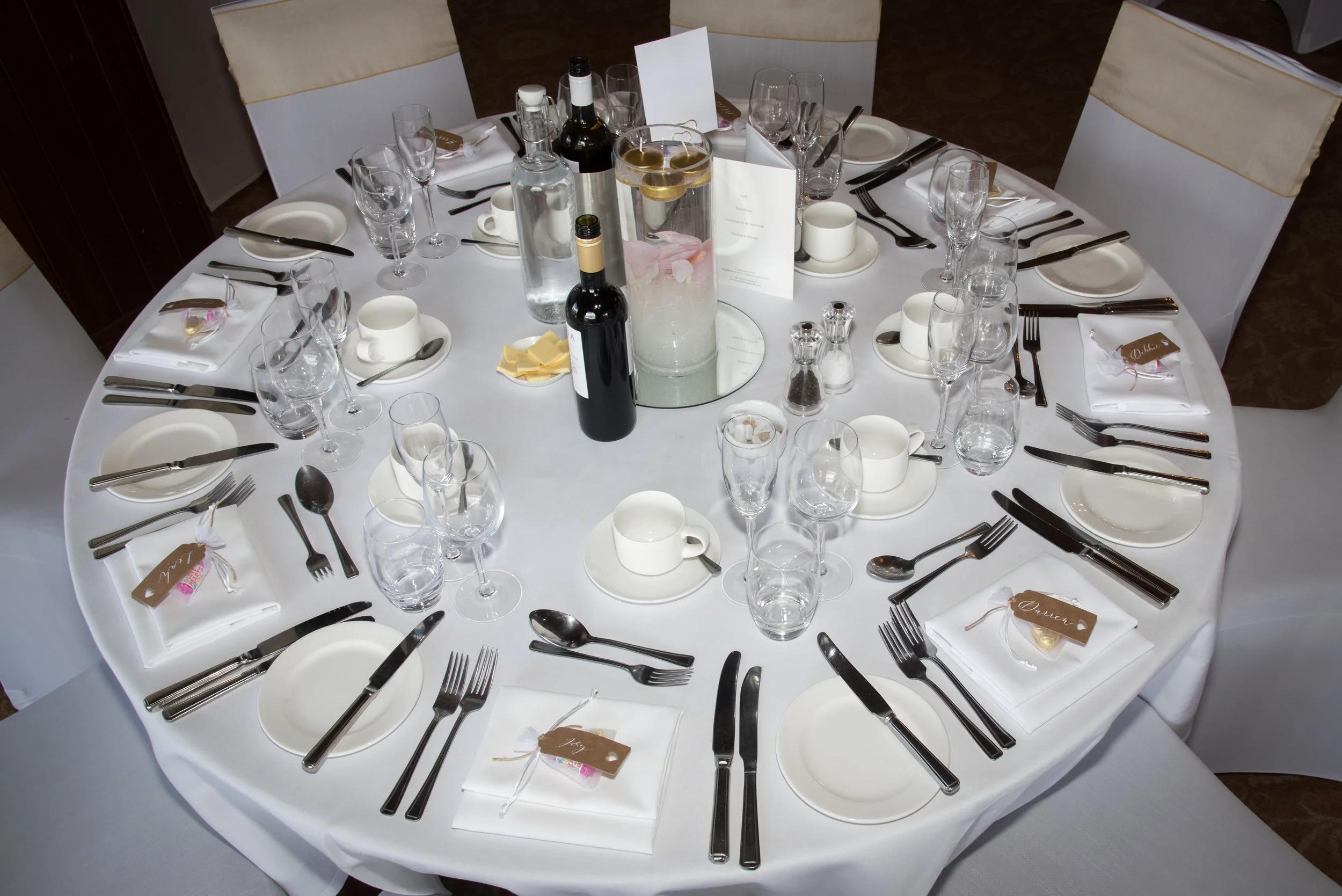 A round banquet table set for a formal meal with white tablecloth, white plates, multiple wine glasses, utensils, and small white cups and saucers. In the center are two bottles of wine, a water pitcher, a glass bowl with floating lemons, and a small