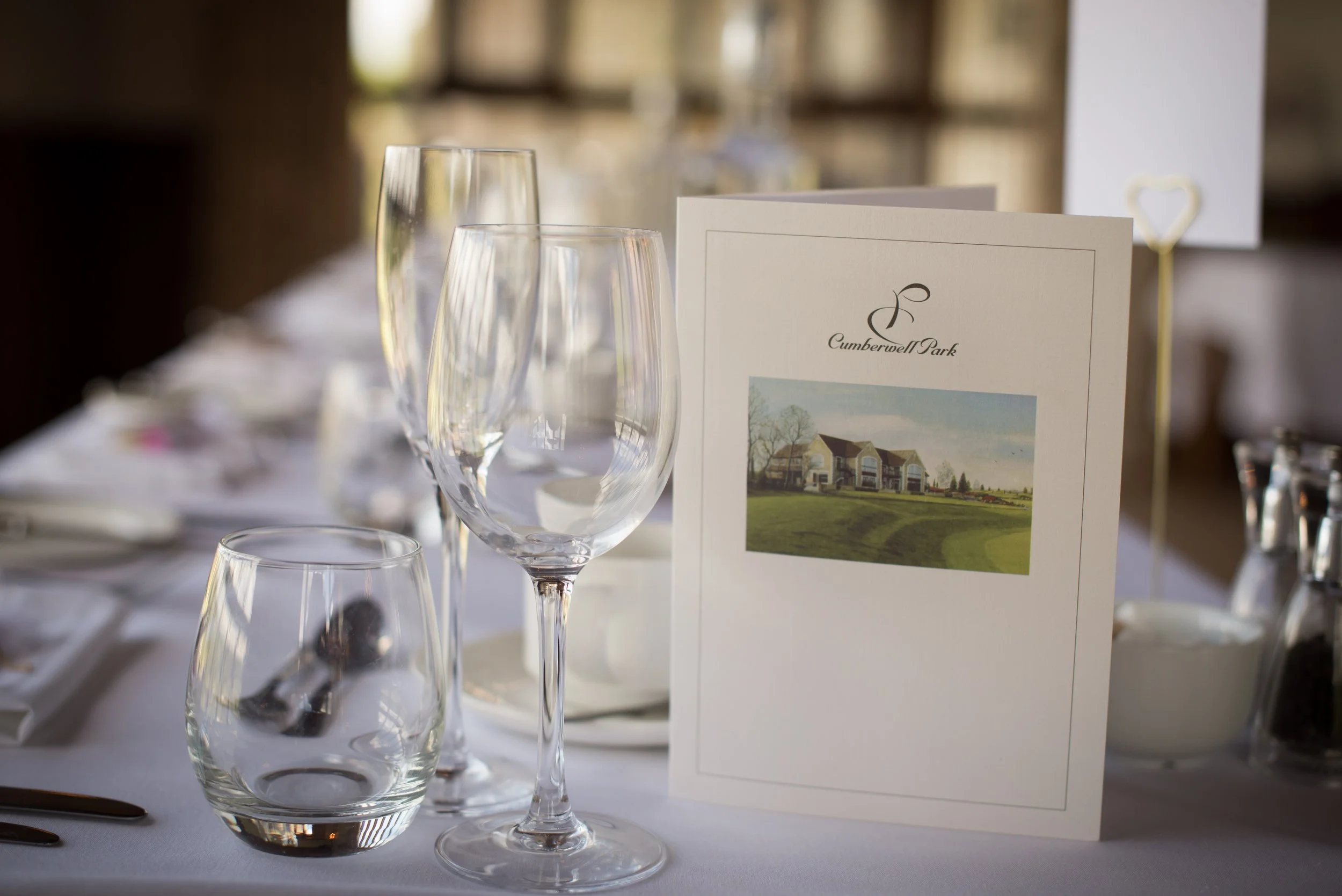 A table set with wine glasses, water glasses, and a white menu with a photo of a house and the text 'Cumberwell Park'.