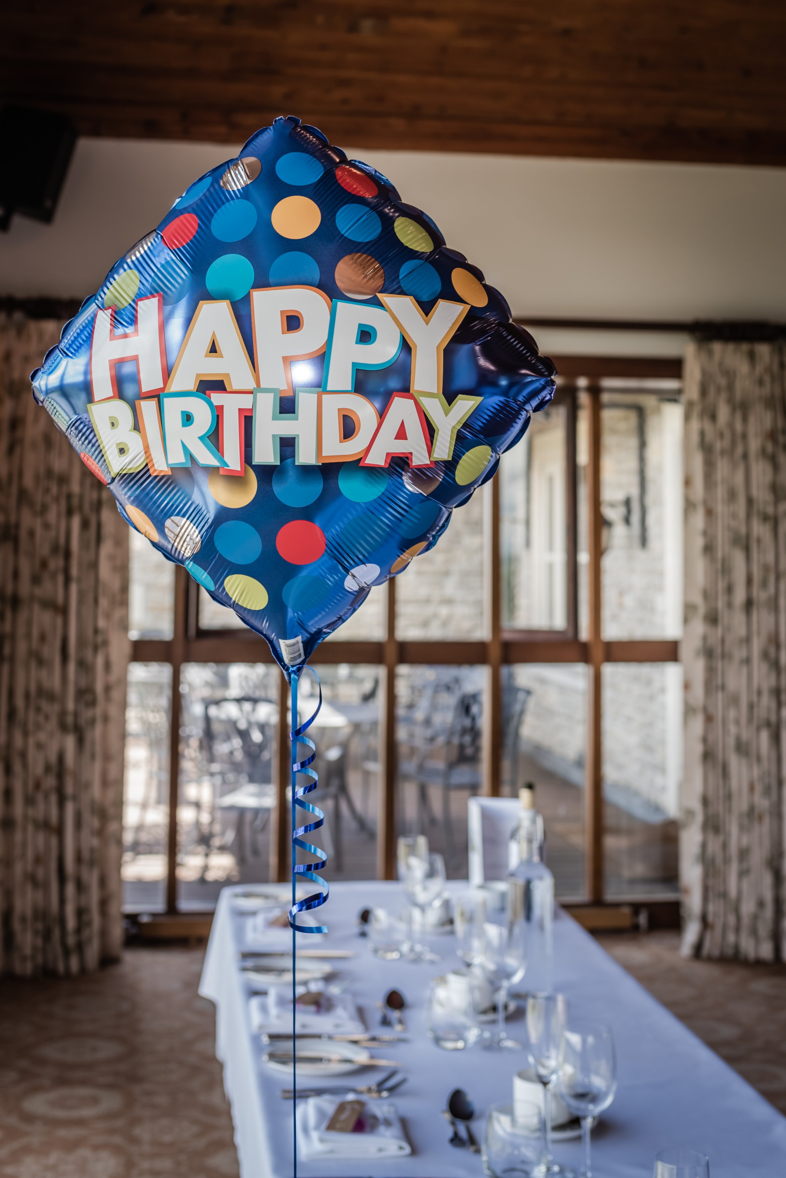 A birthday balloon with colorful polka dots and the message "Happy Birthday" floating over a dining table set for a celebration, in a room with large windows and curtains.