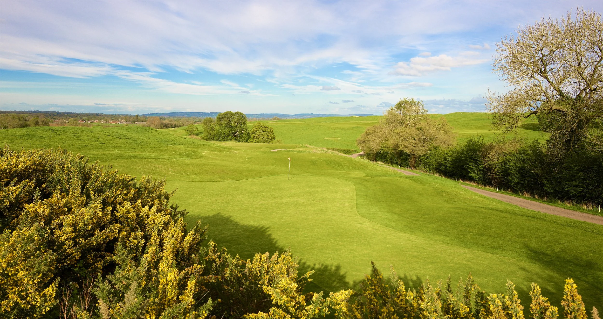 A vibrant green golf course located in a lush, hilly landscape with trees and shrubs, under a partly cloudy sky.