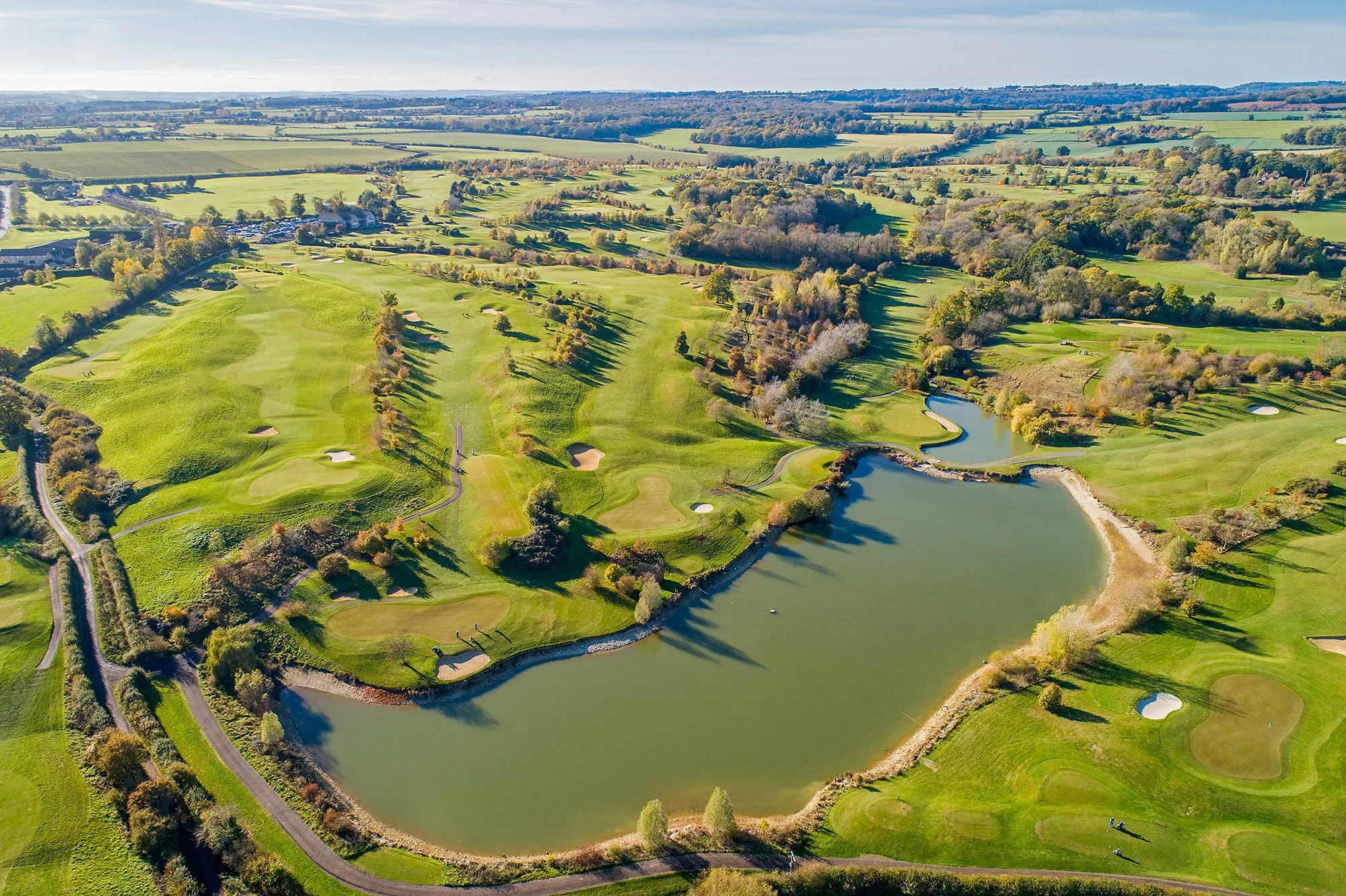 Aerial view of Cumberwell Park golf course with green fairways, sand traps, water hazards, surrounded by trees and open fields under a partly cloudy sky.