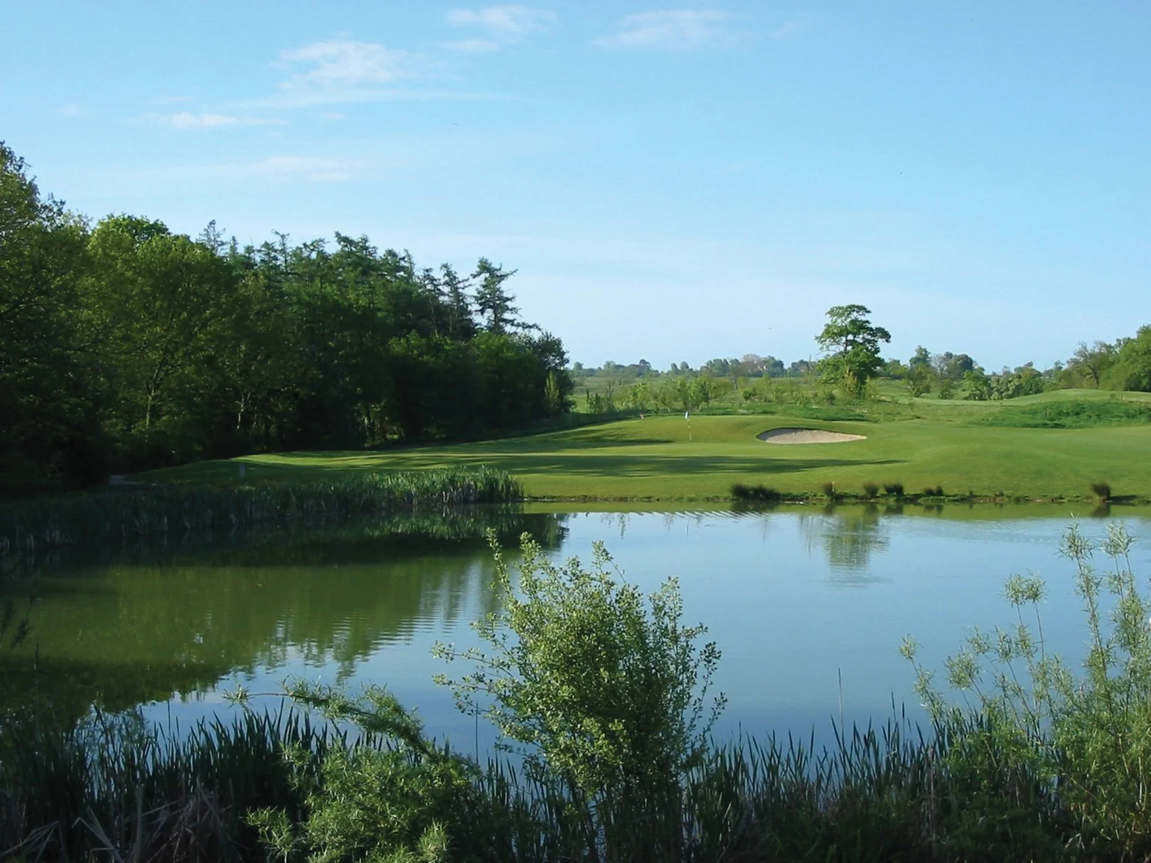 A golf course with a water hazard, trees, and a green with a flagstick in the distance on a clear day.