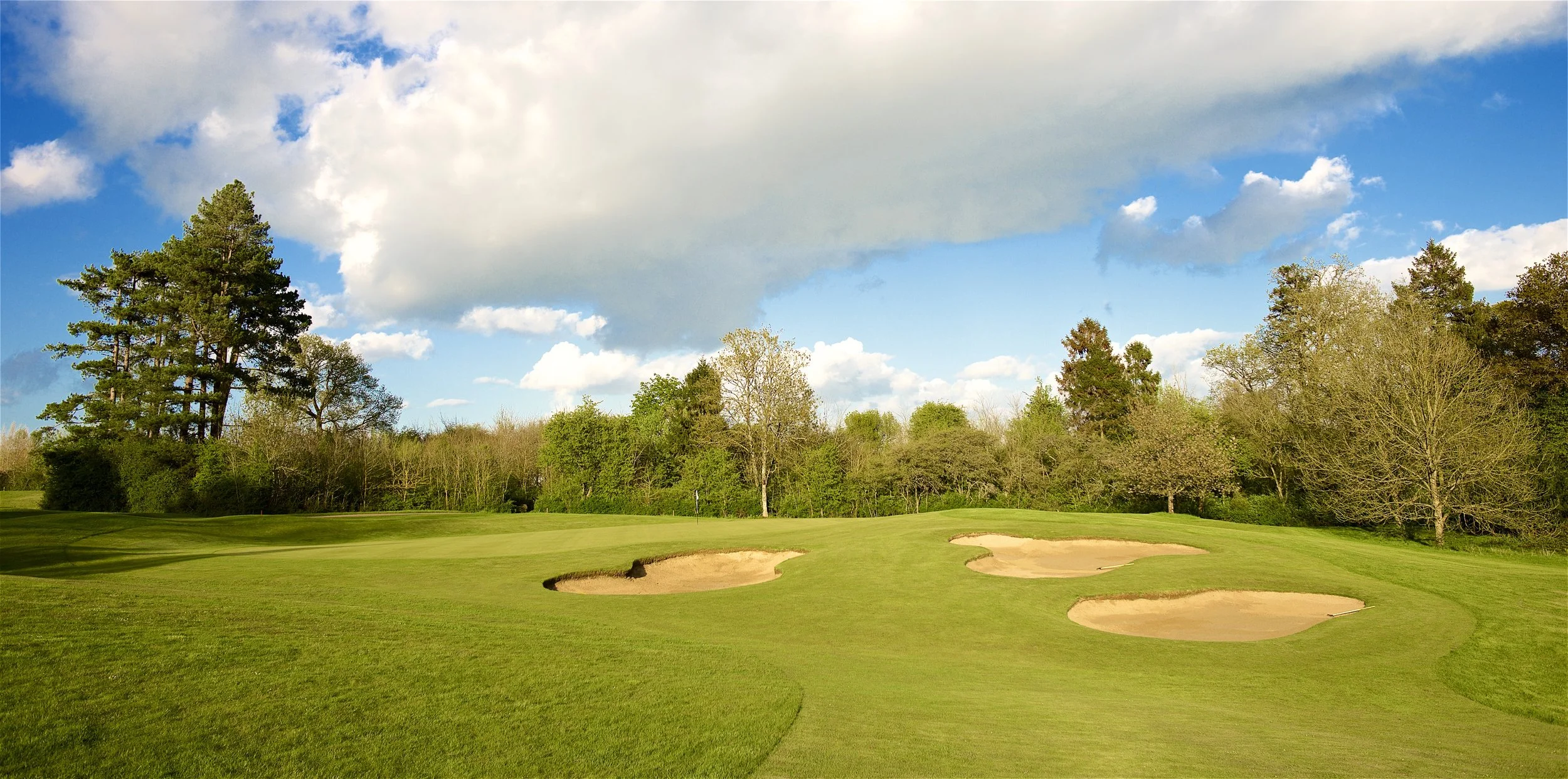 A scenic view of Cumberwell Park golf course with sand bunkers, lush green grass, and a backdrop of trees under a partly cloudy sky.