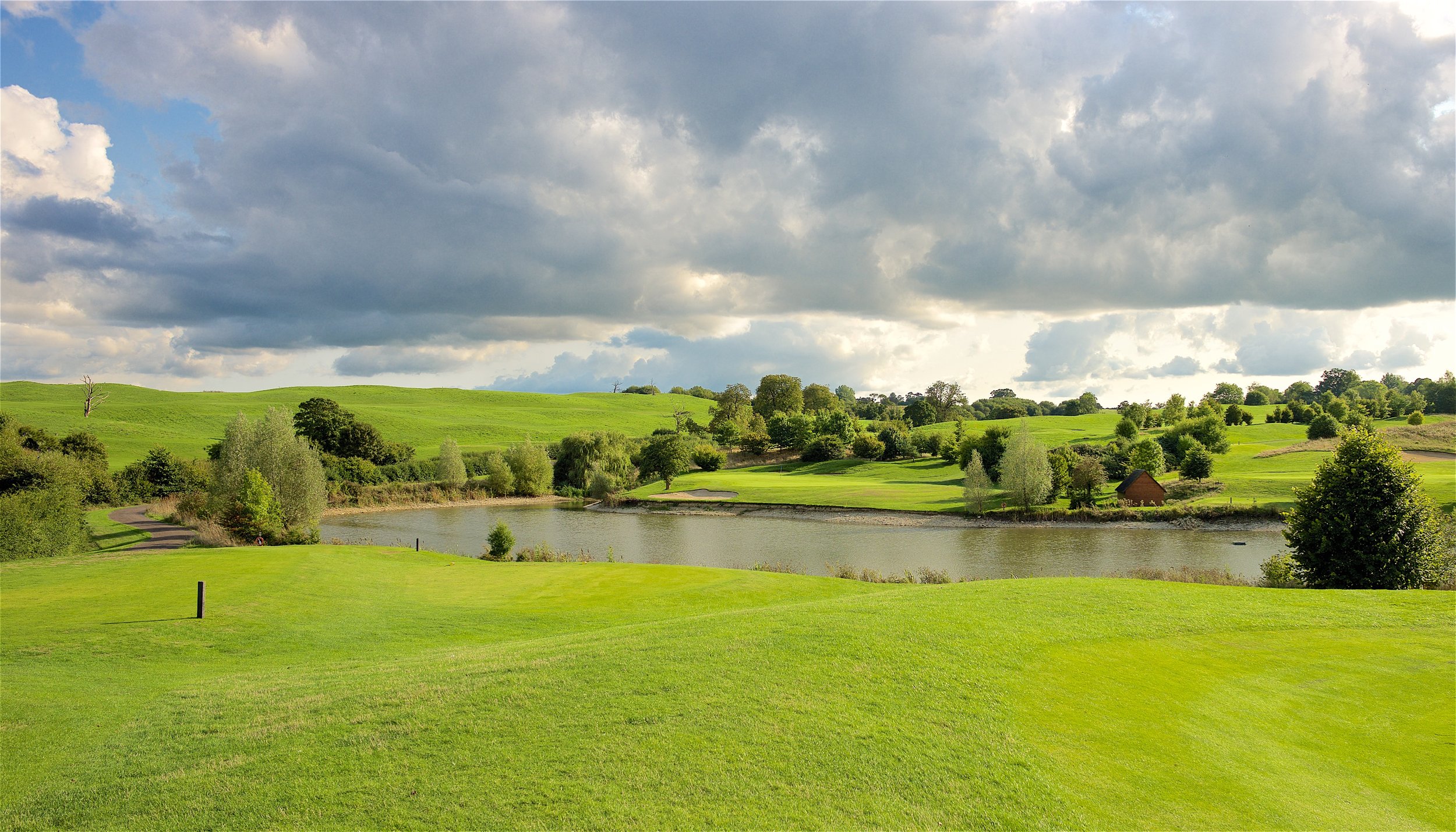Scenic view of a golf course with lush green grass, trees, a body of water, and cloudy sky.
