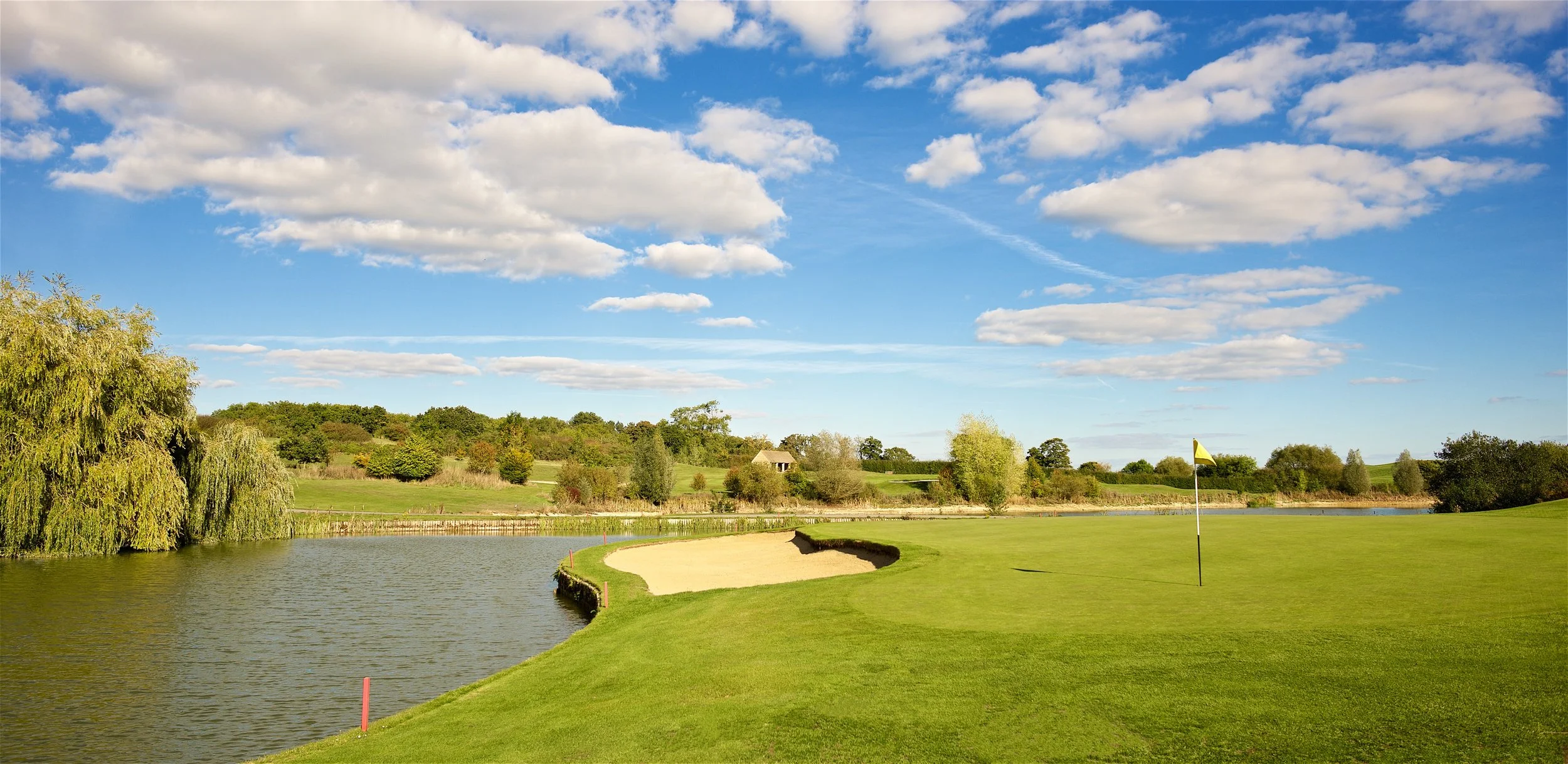 A scenic golf course with a sand trap near a water hazard, green grass, a golf flag, and trees under a partly cloudy sky.