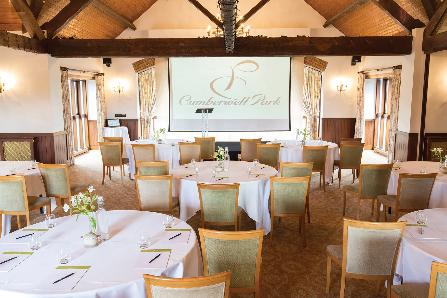 A conference or event room set up with round tables covered in white tablecloths, each with chairs arranged around them. The tables have minimal items, including glasses, notepads, and pens. In the background, there is a large projector screen displaying the logo and name "Cumberwell Park." The room has wooden beams, wall sconces, windows with floral curtains, and a chandelier hanging from the ceiling.