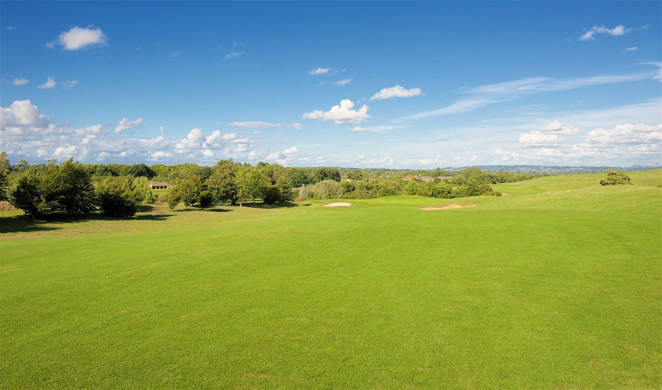 A scenic view of a golf course with green grass, sand bunkers, trees, and a clear blue sky with scattered clouds.