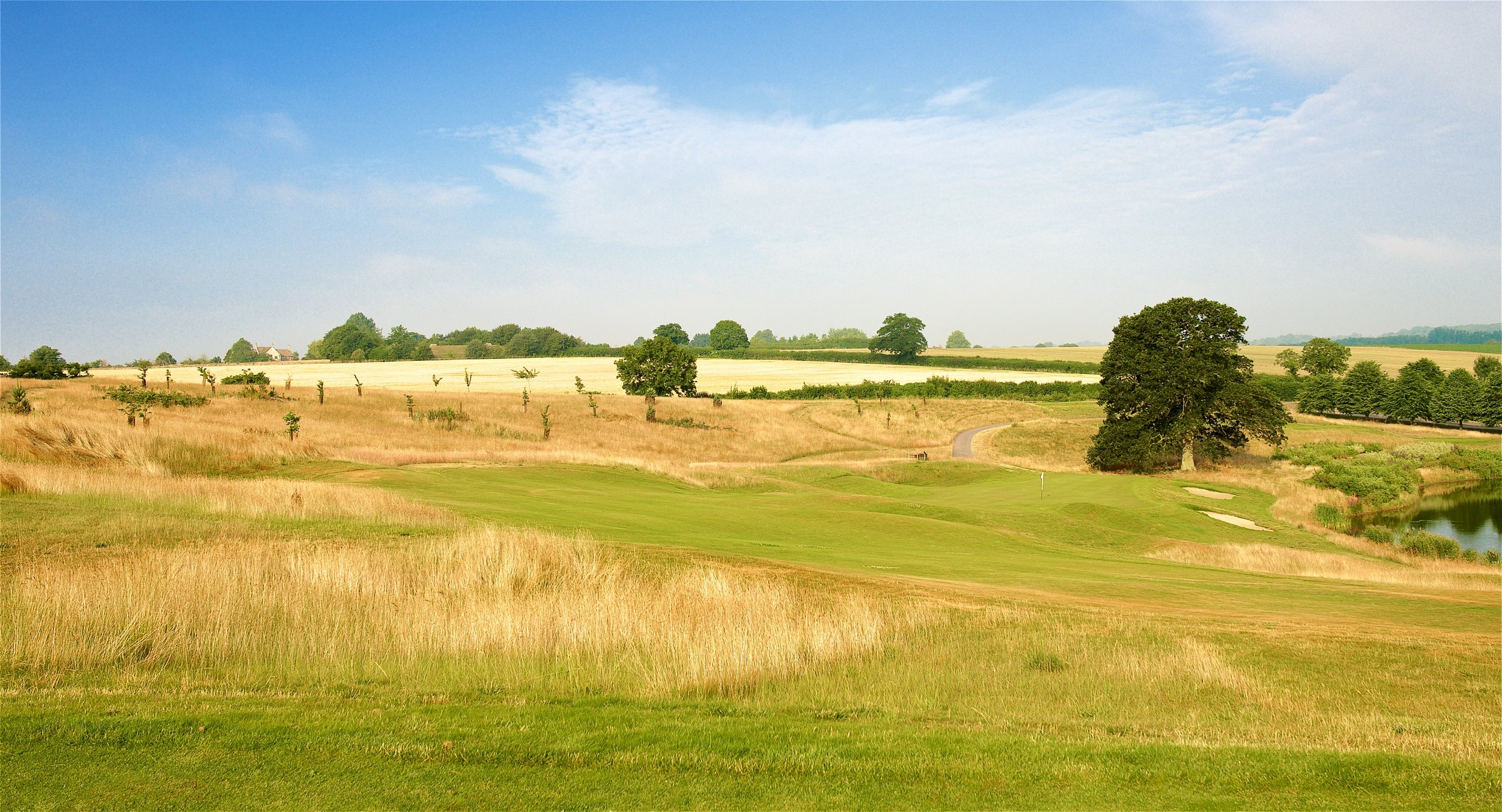 A scenic landscape of a golf course with green fairways, sand bunkers, trees, and a small pond, set against rolling fields and a bright blue sky with scattered clouds.