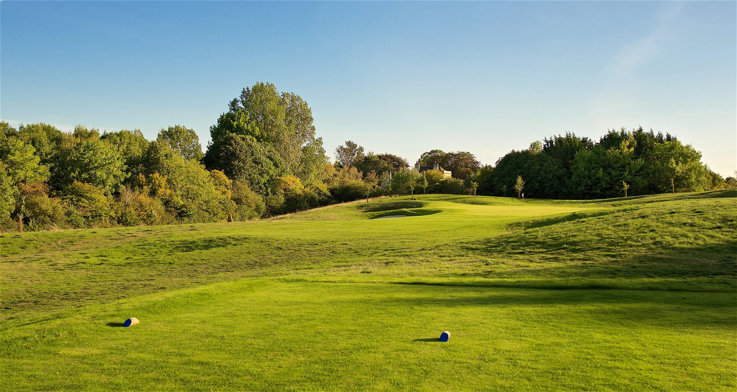 A golf course with a green fairway, sand bunkers, and a wooded background under a clear blue sky.