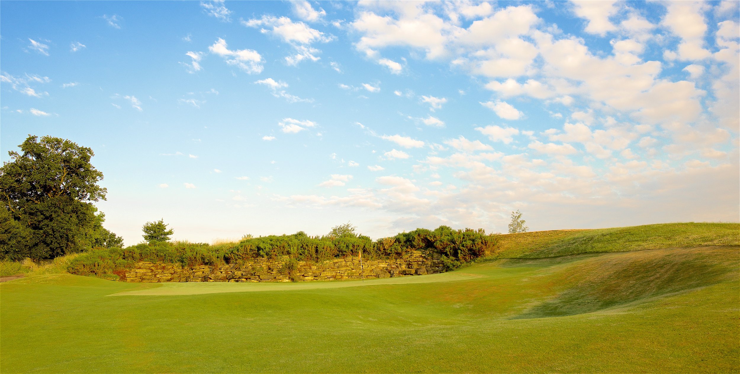 A golf course with green grass, a stone wall, trees, and a blue sky with scattered clouds.