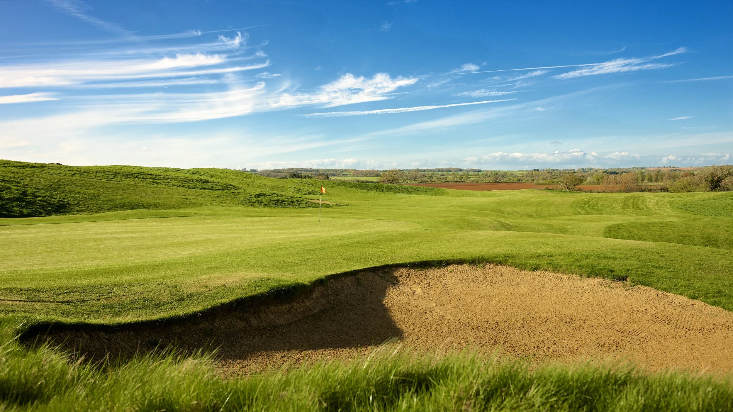 A golf course with a sand bunker in the foreground, lush green grass, a flag on a hole, and expansive open landscape under a bright blue sky with streaks of white clouds.