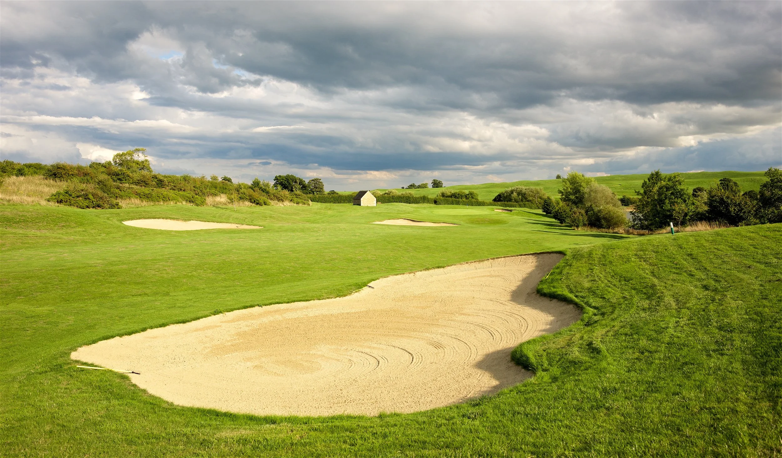 A golf course with green grass and sand bunkers, surrounded by trees and a cloudy sky.