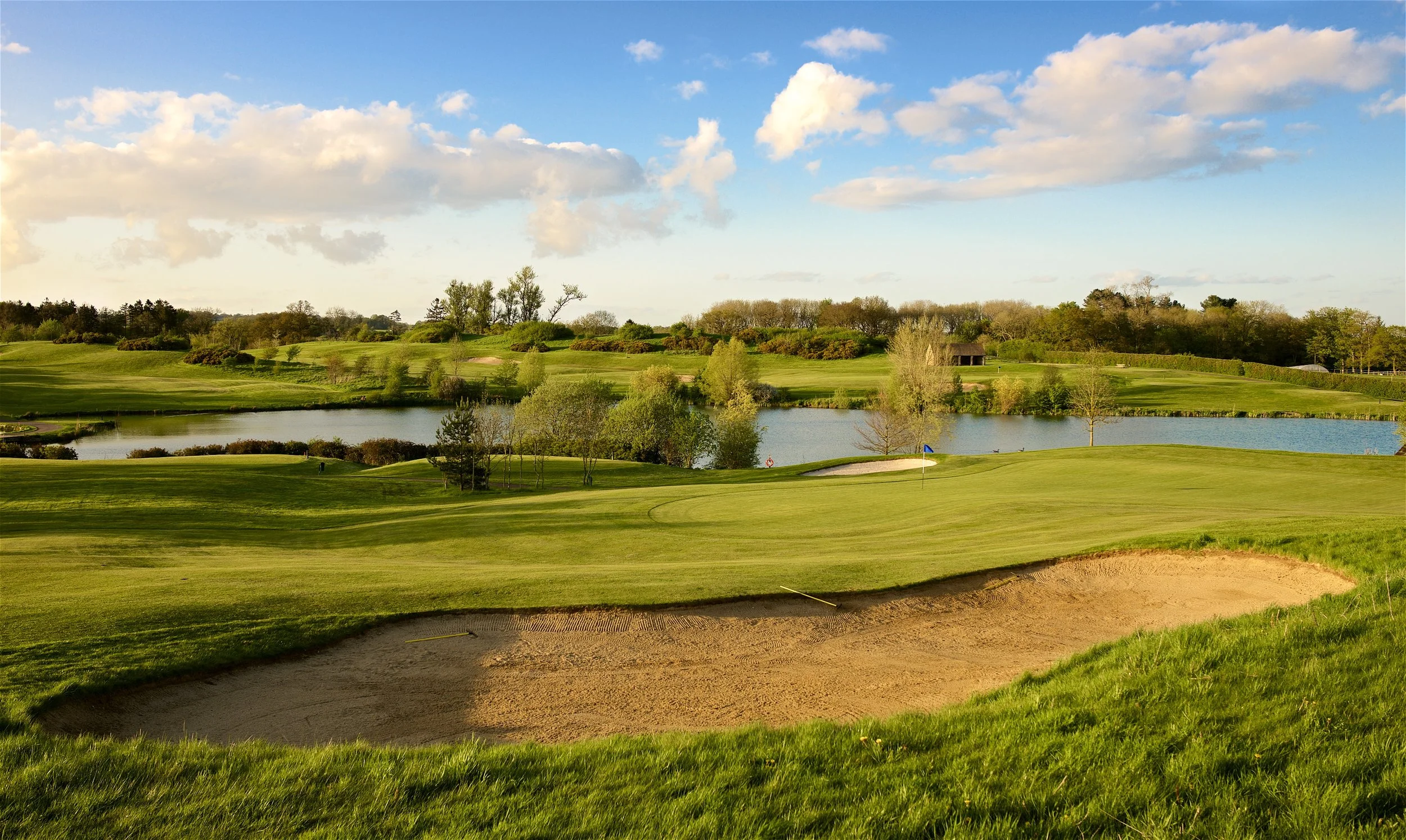 A scenic view of a golf course with green fairways, a pond, sand traps, and trees under a blue sky with fluffy clouds.