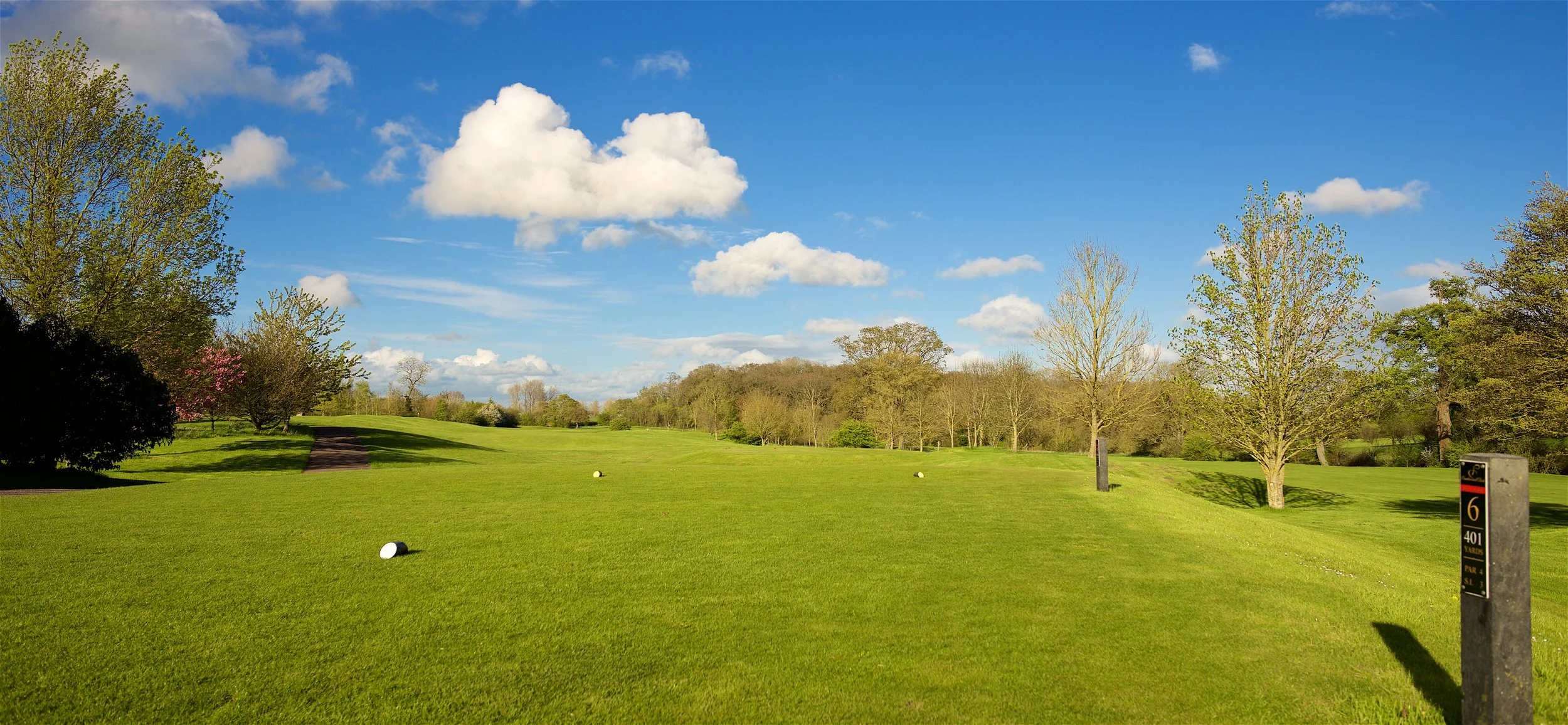 A golf course with lush green grass, several trees, and a bright blue sky with white clouds. The course has a sign indicating the 6th hole, 401 yards, par 4.
