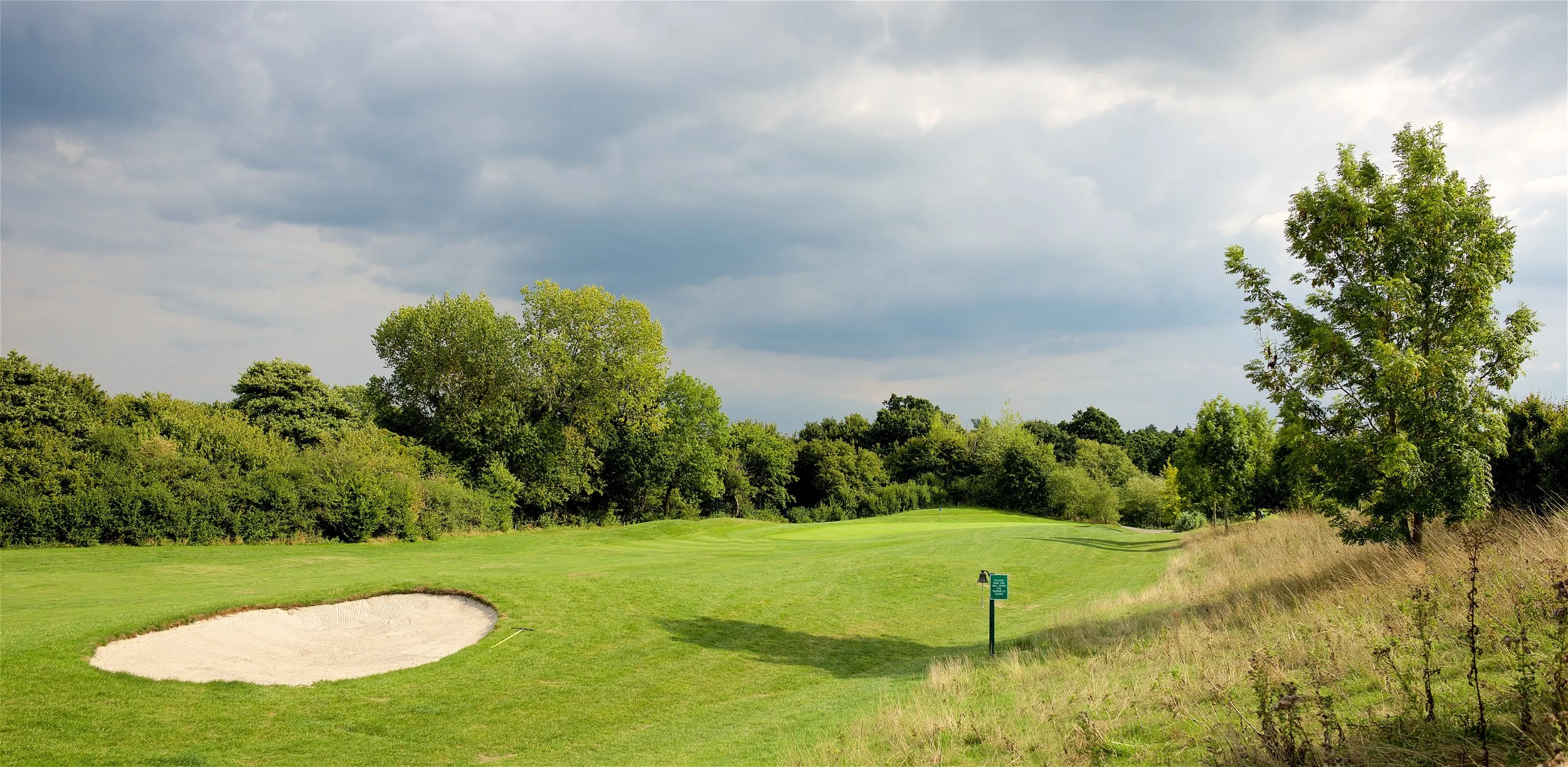 A golf course with a sand trap in the foreground, lush green grass, trees in the background, and a cloudy sky overhead.