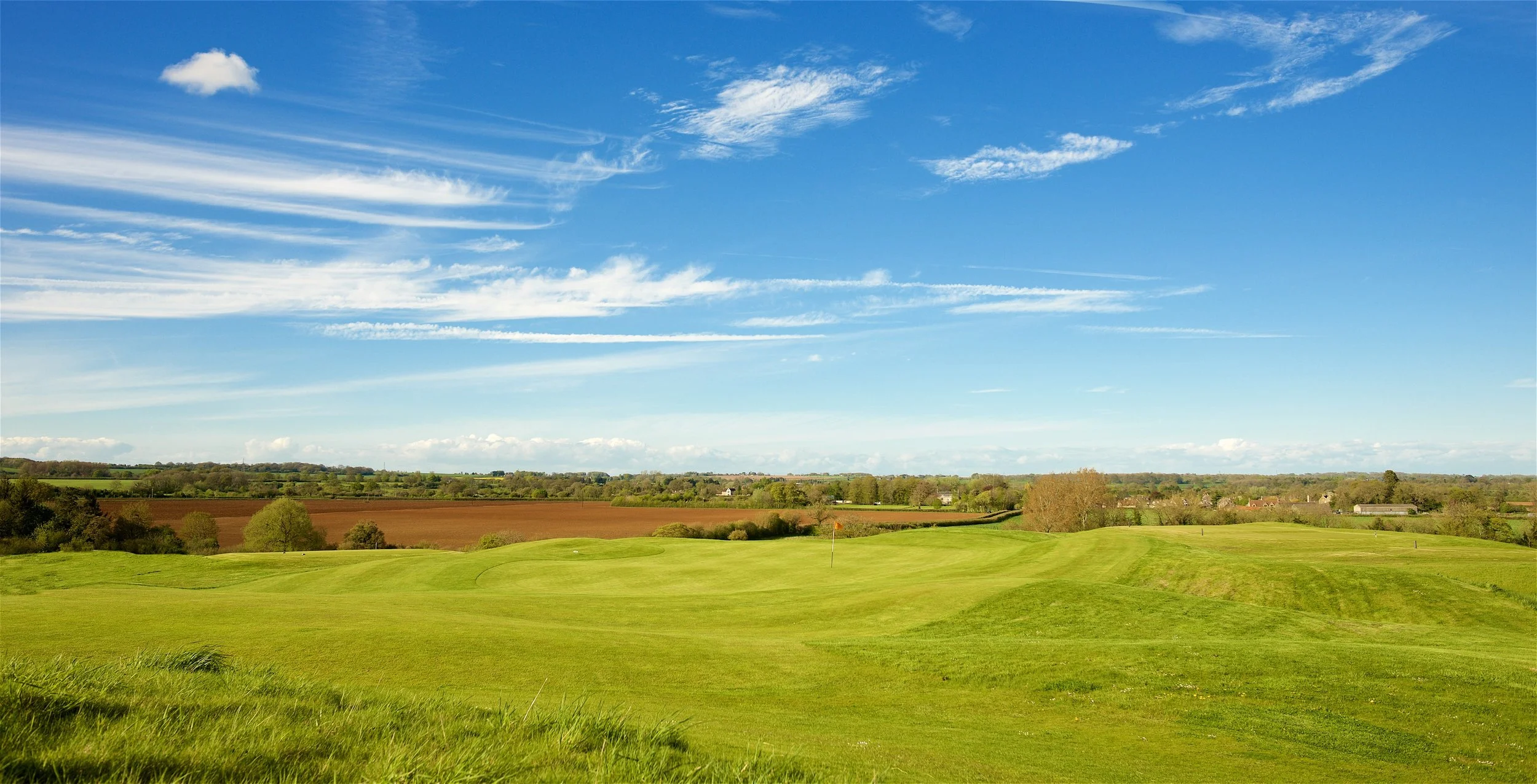 A golf course with a flag on the green, green grass, rolling landscape, and a partly cloudy blue sky.
