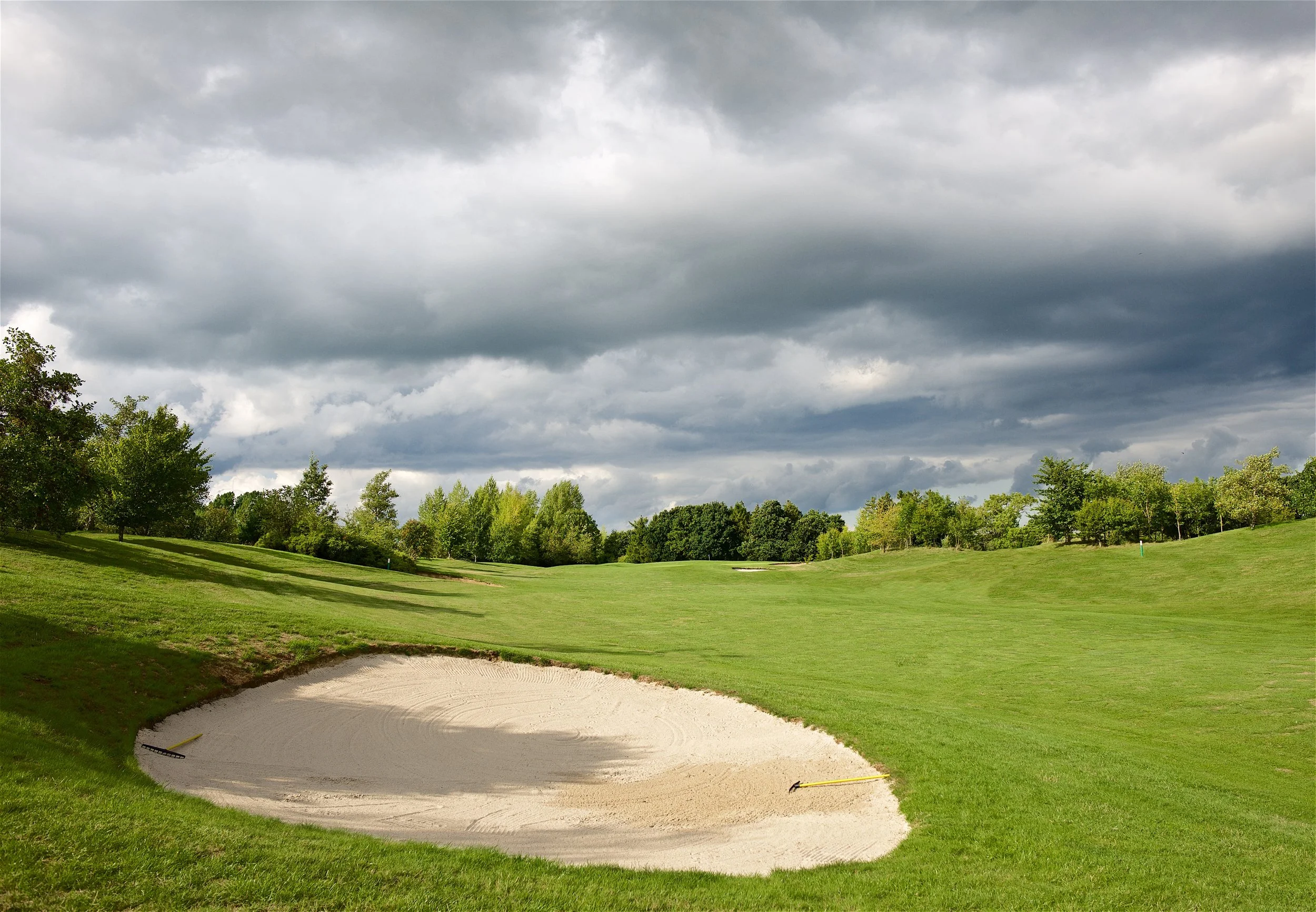 A golf course with a sand trap bunker in the foreground, lush green grass, and trees in the background under a cloudy sky.