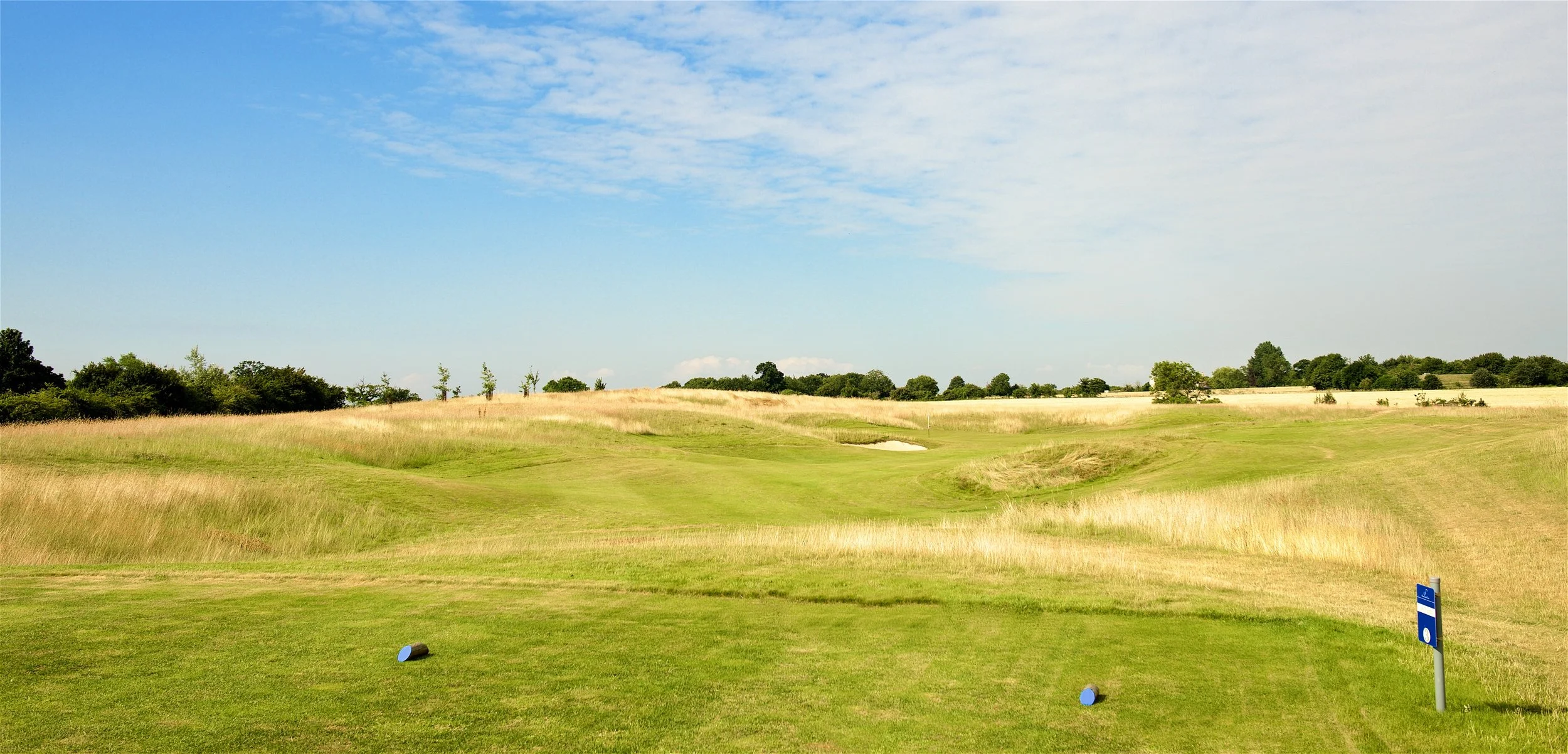 A golf course with green grass, sand traps, and a flag in the distance under a partly cloudy blue sky.