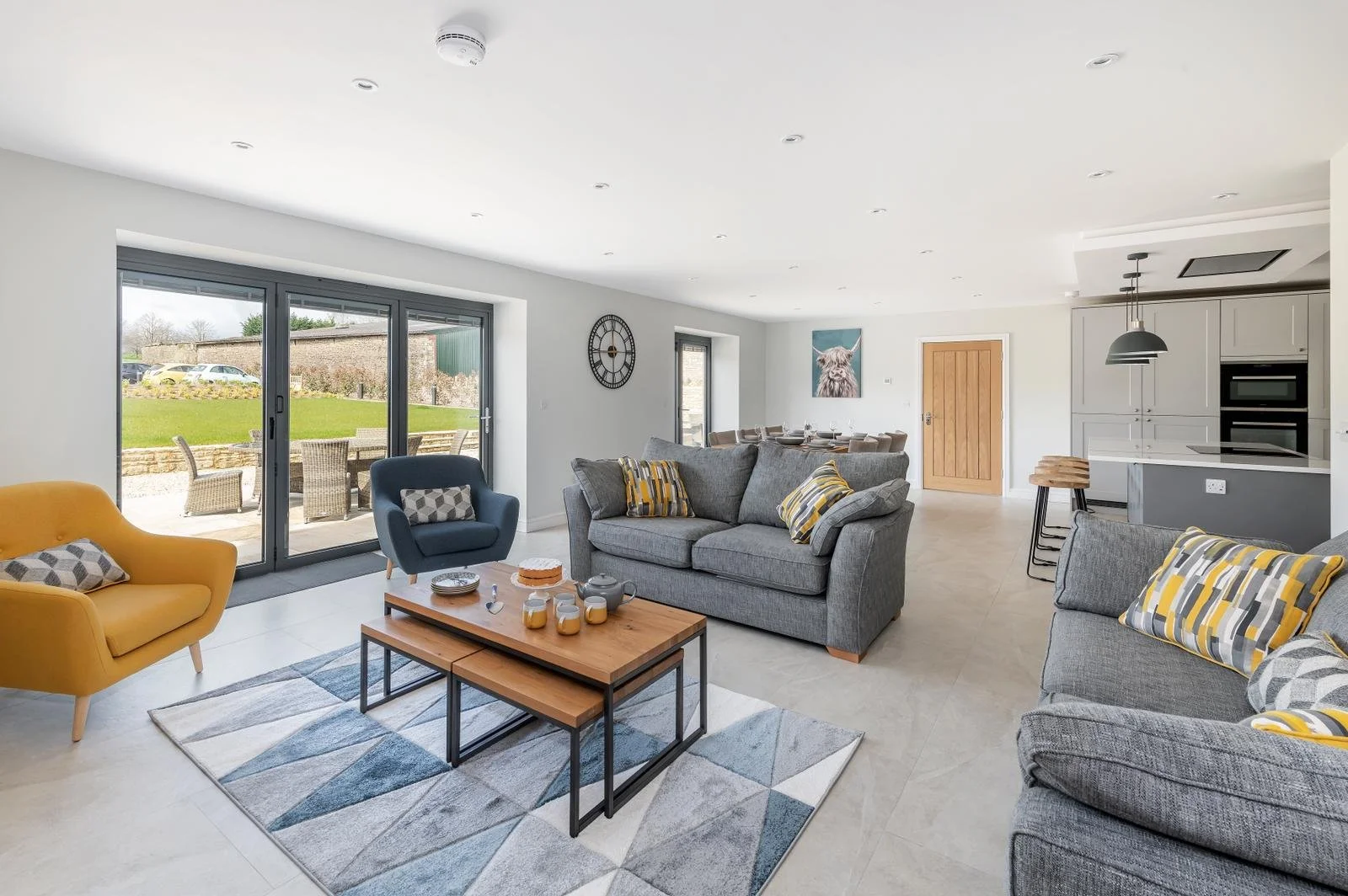 Interior of a modern, spacious living room with gray and yellow furniture, a wooden coffee table, a large gray sofa with striped pillows, a blue armchair, a yellow armchair, and sliding glass doors leading outside to a patio with outdoor chairs and a green lawn.