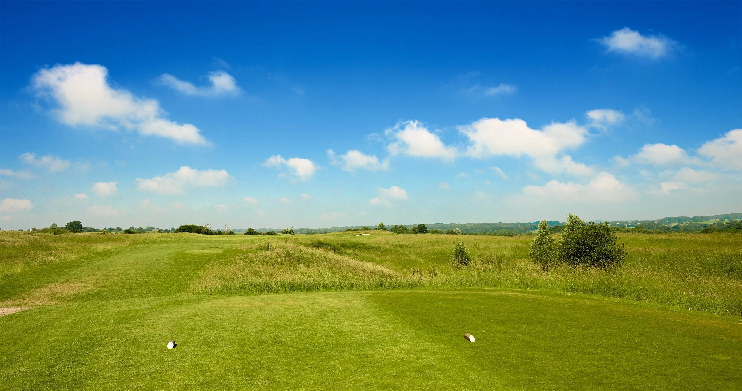 A golf course with lush green grass, trees, and a path in the foreground under a partly cloudy blue sky.