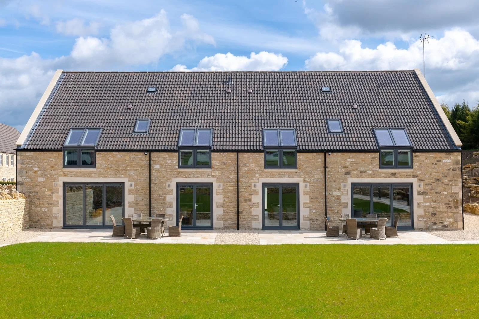 Stay and Play at Cumberwell Park golf course. A modern multi-story house with brick exterior, multiple large windows, and a tiled roof, surrounded by a well-maintained lawn and patio furniture.