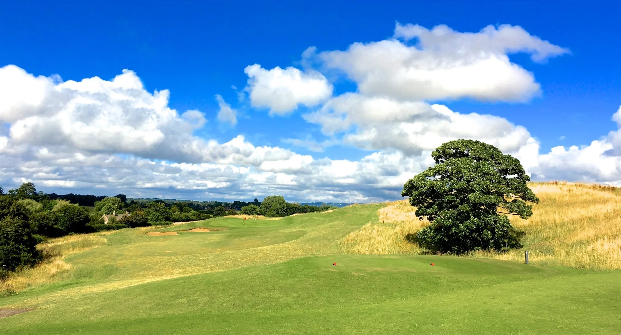 A bright blue sky with scattered fluffy white clouds over a golf course. The course has neatly trimmed green grass, sand bunkers, and a large leafy tree on the right side. Rolling hills and greenery extend into the distance.