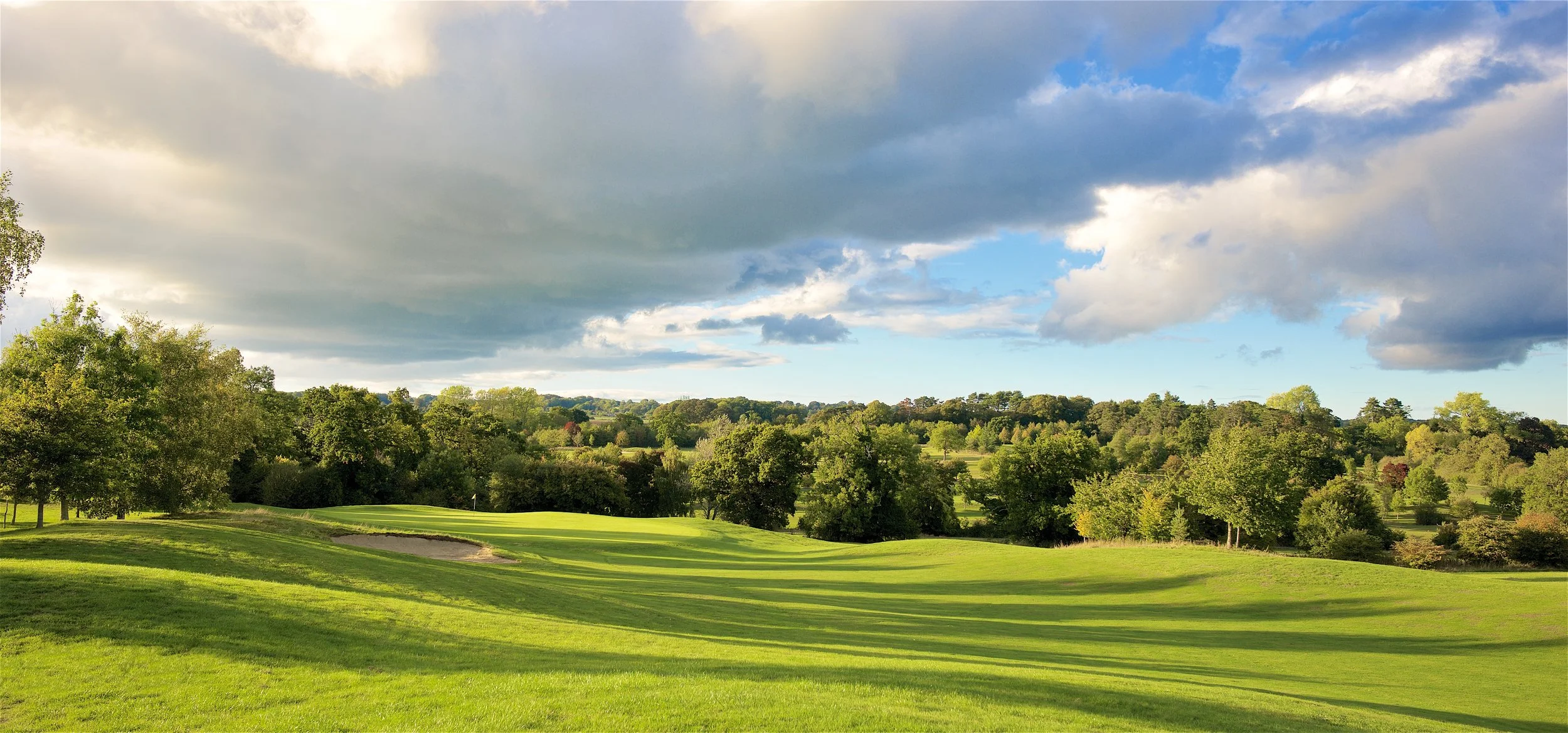 A lush green golf course in southern England with a sand trap in the foreground, surrounded by dense trees under a partly cloudy sky.