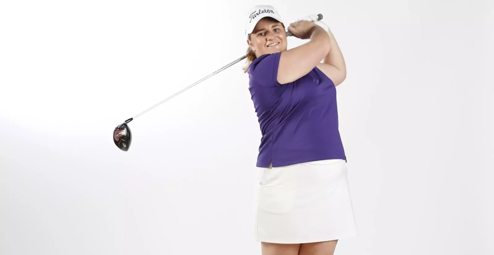 A woman in a purple golf shirt and white skirt holding a golf club, smiling, against a plain white background.