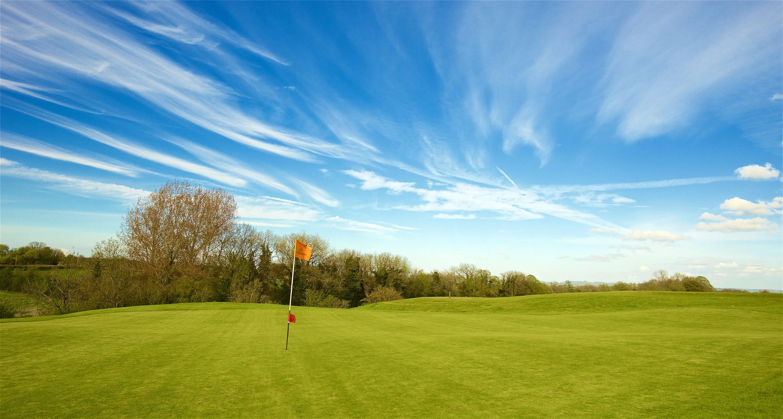 A golf course green with a flagstick, surrounded by trees and a blue sky with wispy clouds.