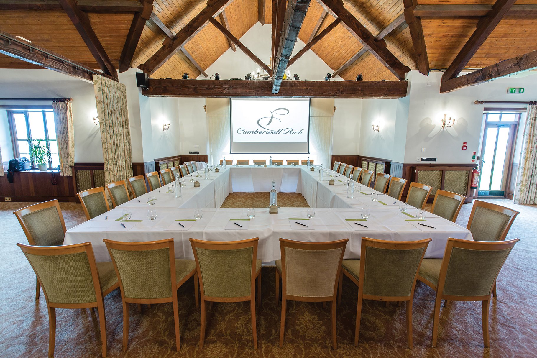 Meeting room with U-shaped white table, chairs, large screen displaying 'Cumberwell Park', wooden beams on vaulted ceiling, windows with floral curtains, and a carpeted floor.