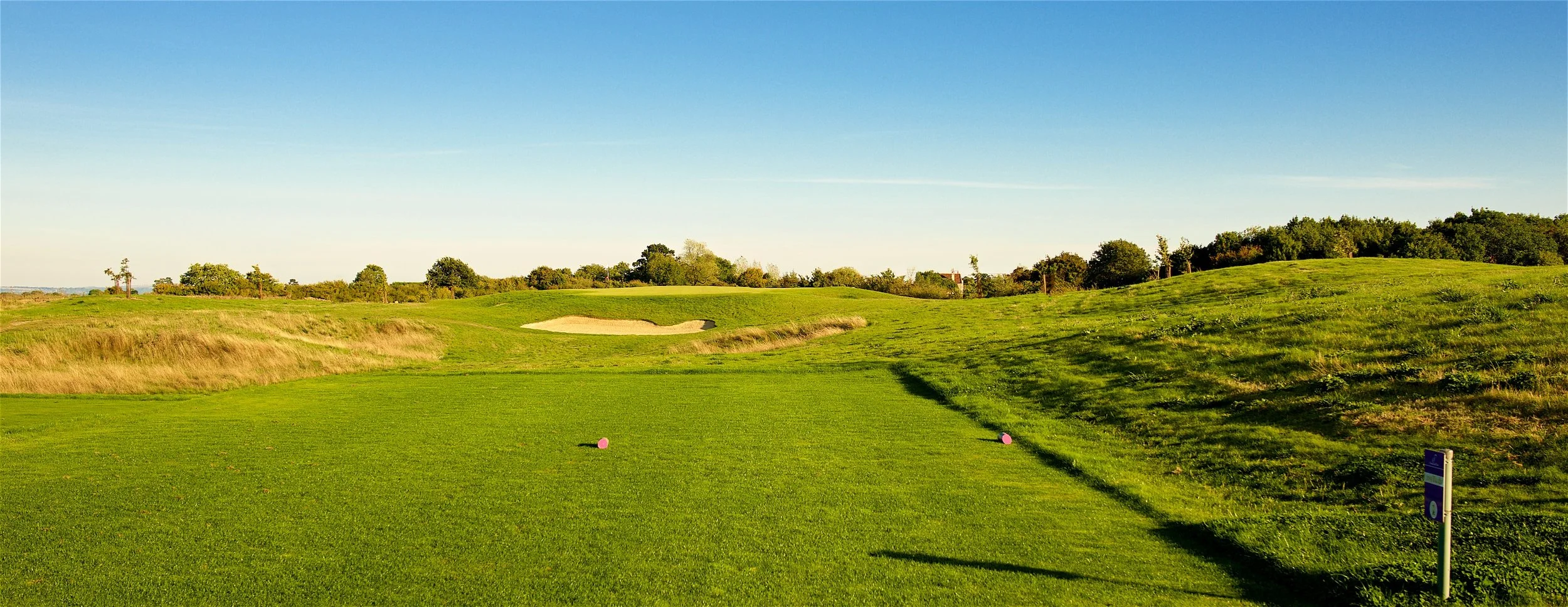 A view of a golf course with a green, sand trap, and tee markers under a clear blue sky.
