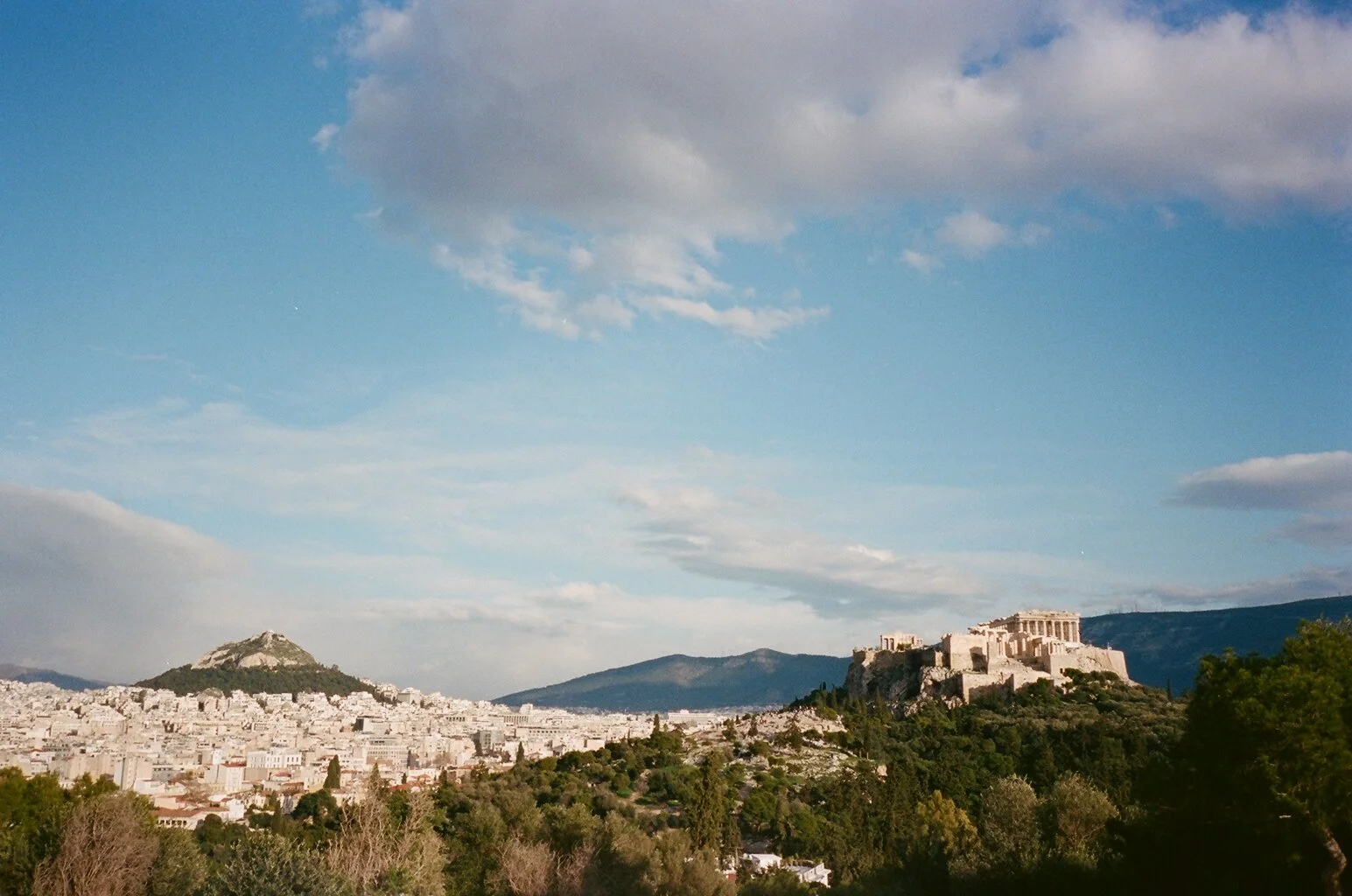 Blick auf die Akropolis in Athen, Griechenland, umgeben von Stadt und Hügeln bei sonnigem Himmel.