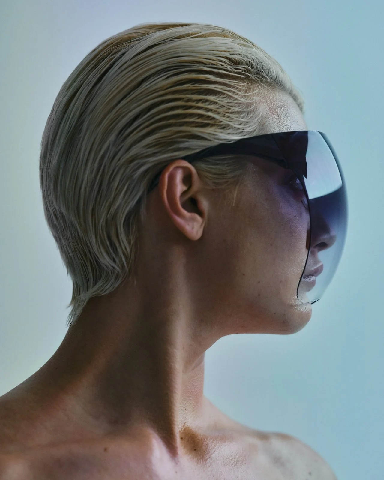 Porträt einer Frau mit blonden, nach hinten geglätteten Haaren, trägt eine große schwarze Sonnenbrille, nur Kopf und obere Brust sichtbar.
