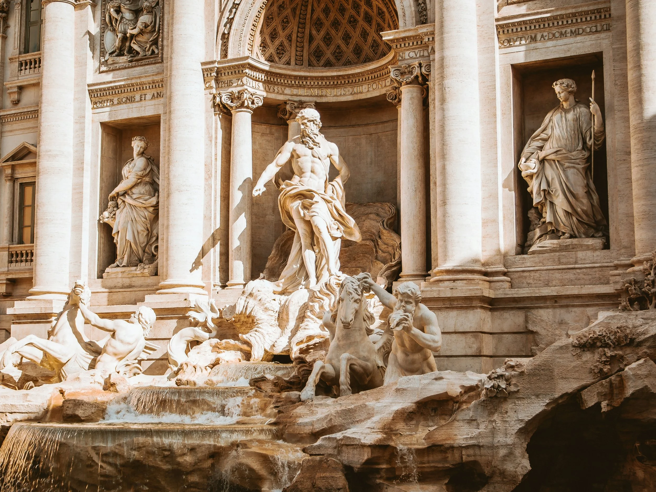 Barockbrunnen mit Skulpturen aus weißem Stein, zentrale Figur ist ein männlicher Gott mit Bart, umgeben von geflügelten Meeresgöttern und Wasserfällen