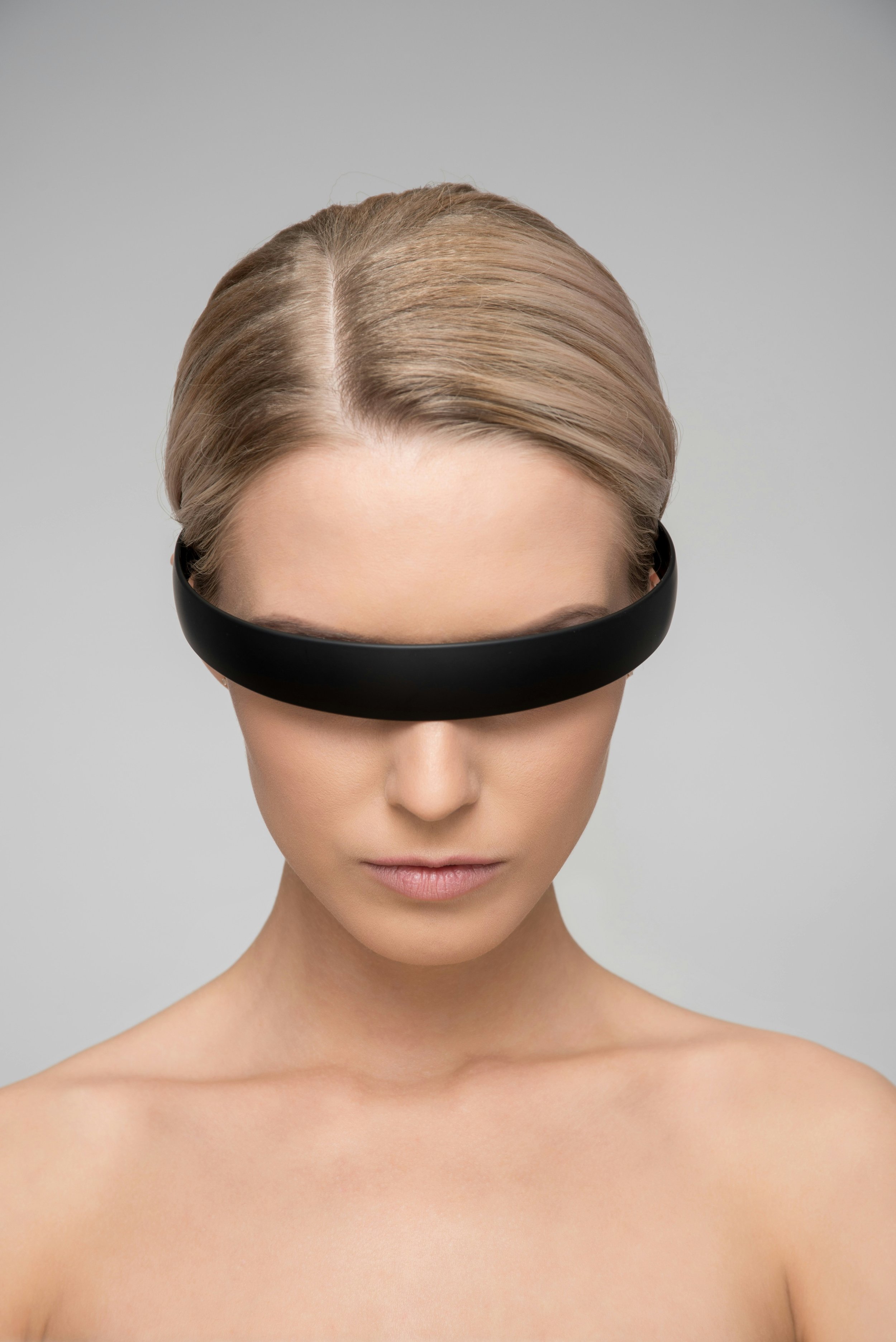Nahaufnahme eines Frauenkopfes mit blondem Haar, der Blick ist nach unten gerichtet, trägt eine schwarze Augenmaske, kein Schmuck sichtbar, nackter Oberkörper, helle Haut, neutraler Hintergrund.