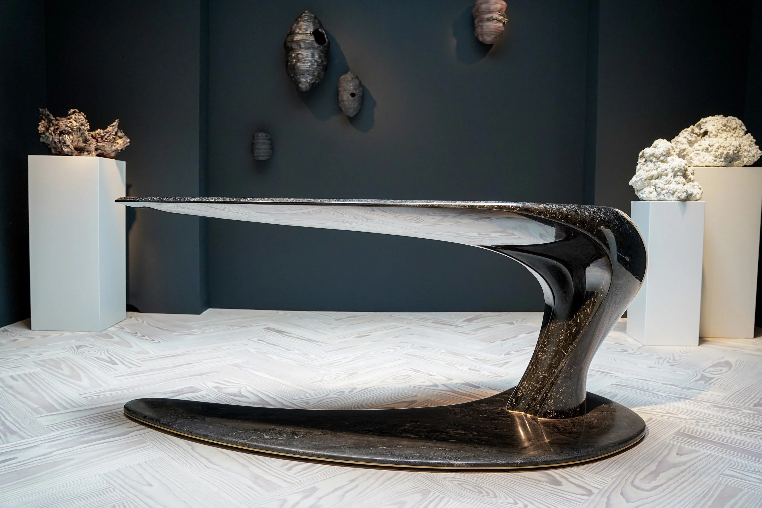 Modern black sculptural table on a light wooden floor, with textured stone artworks mounted on a dark wall and placed on white pedestals.